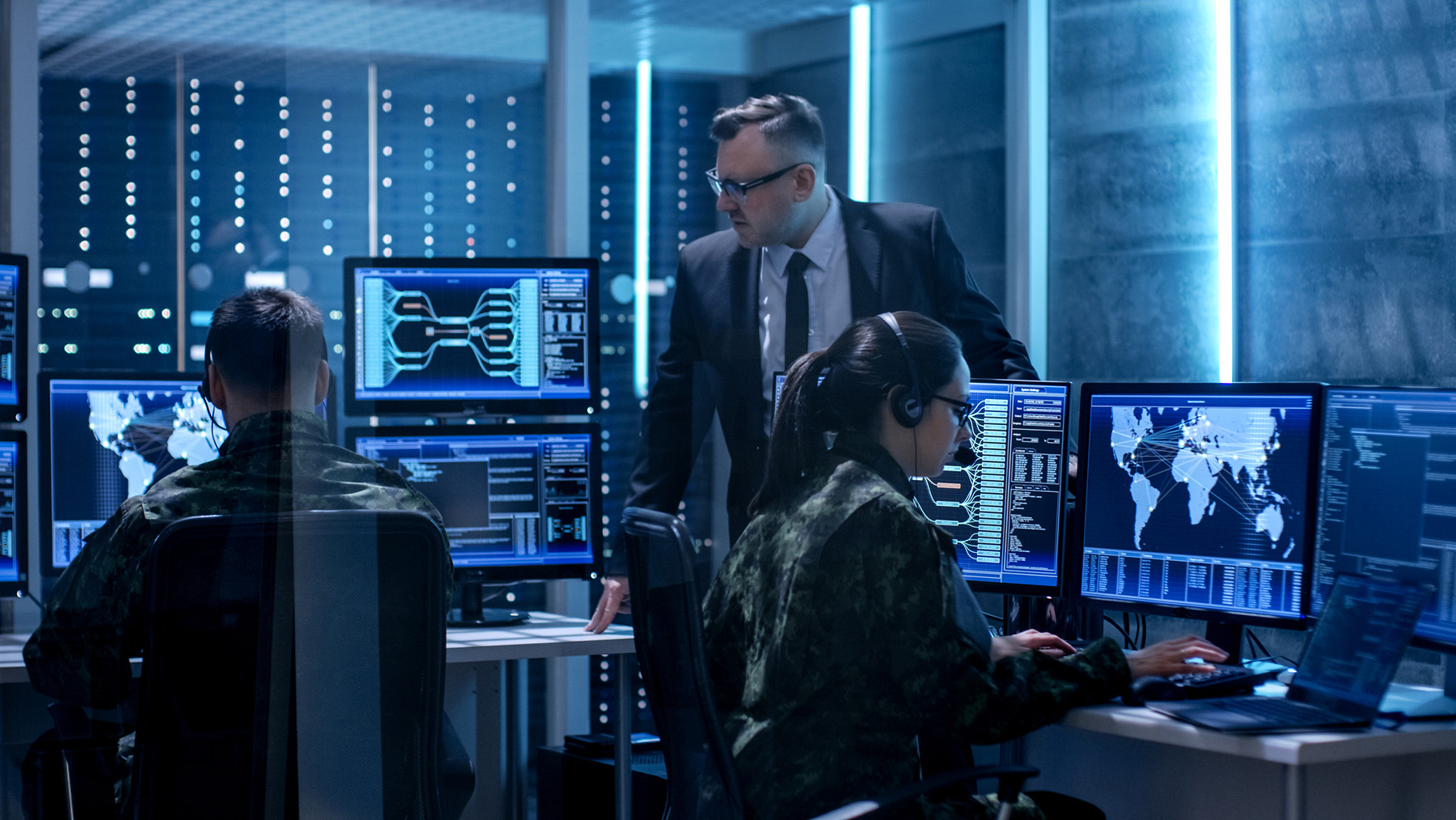 Two military personnel monitoring global cyber networks on multiple computer screens while a man in a suit observes in a high-tech command center.