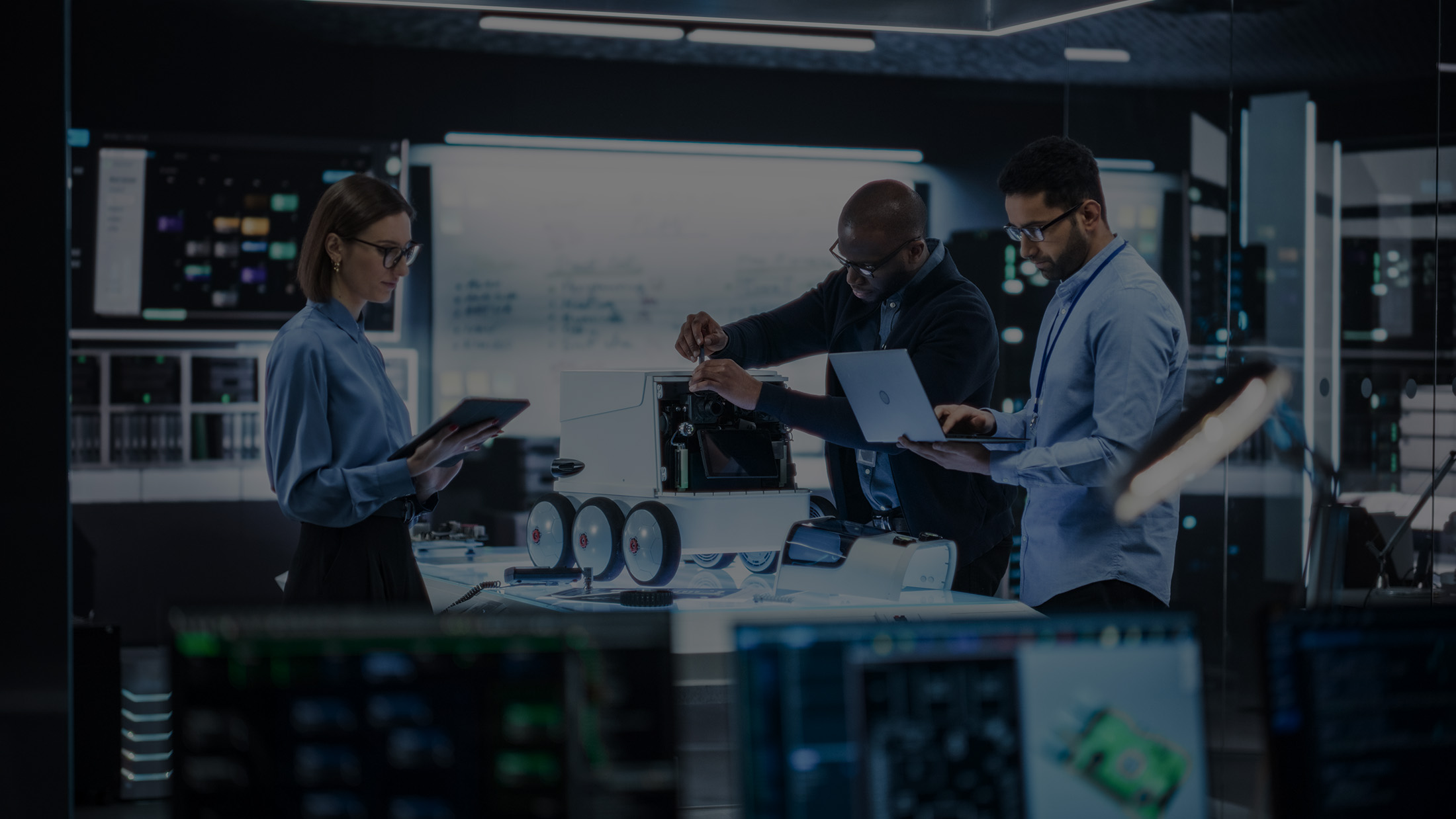 Three engineers working on a wheeled robotic device in a high-tech lab with computers and monitors.