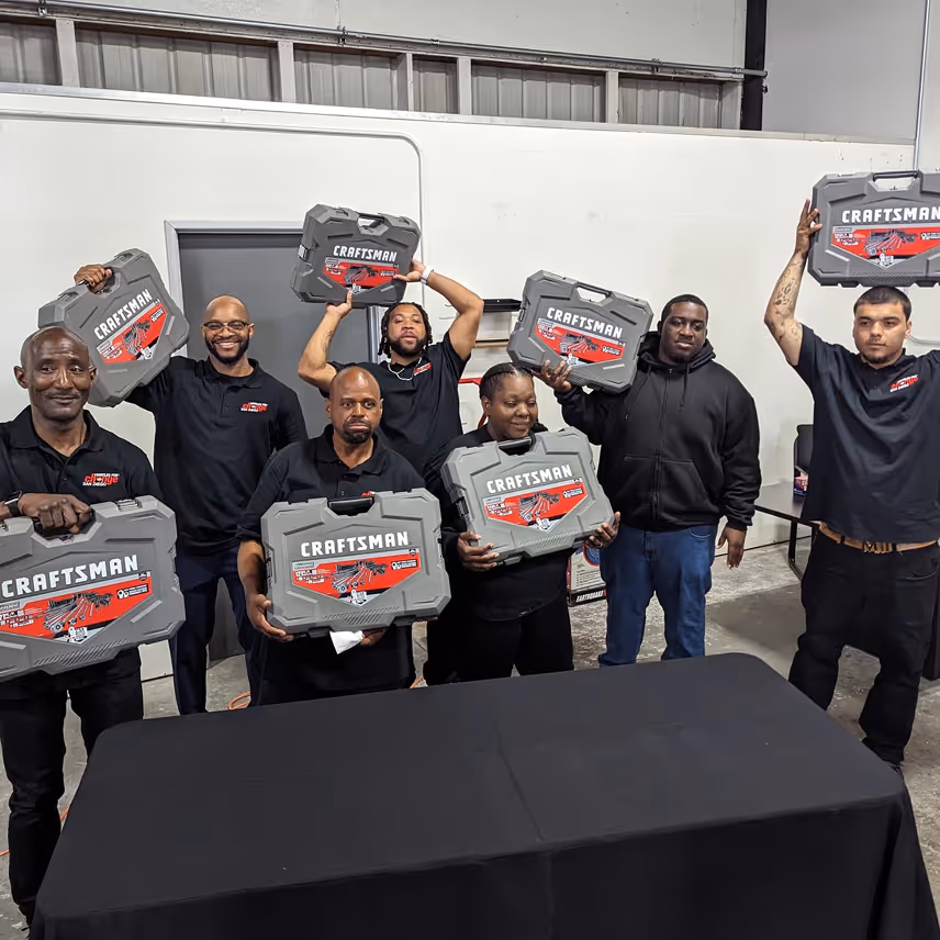 Seven people standing indoors holding gray Craftsman toolkits, some raised above their heads, smiling and posing behind a black-covered table.