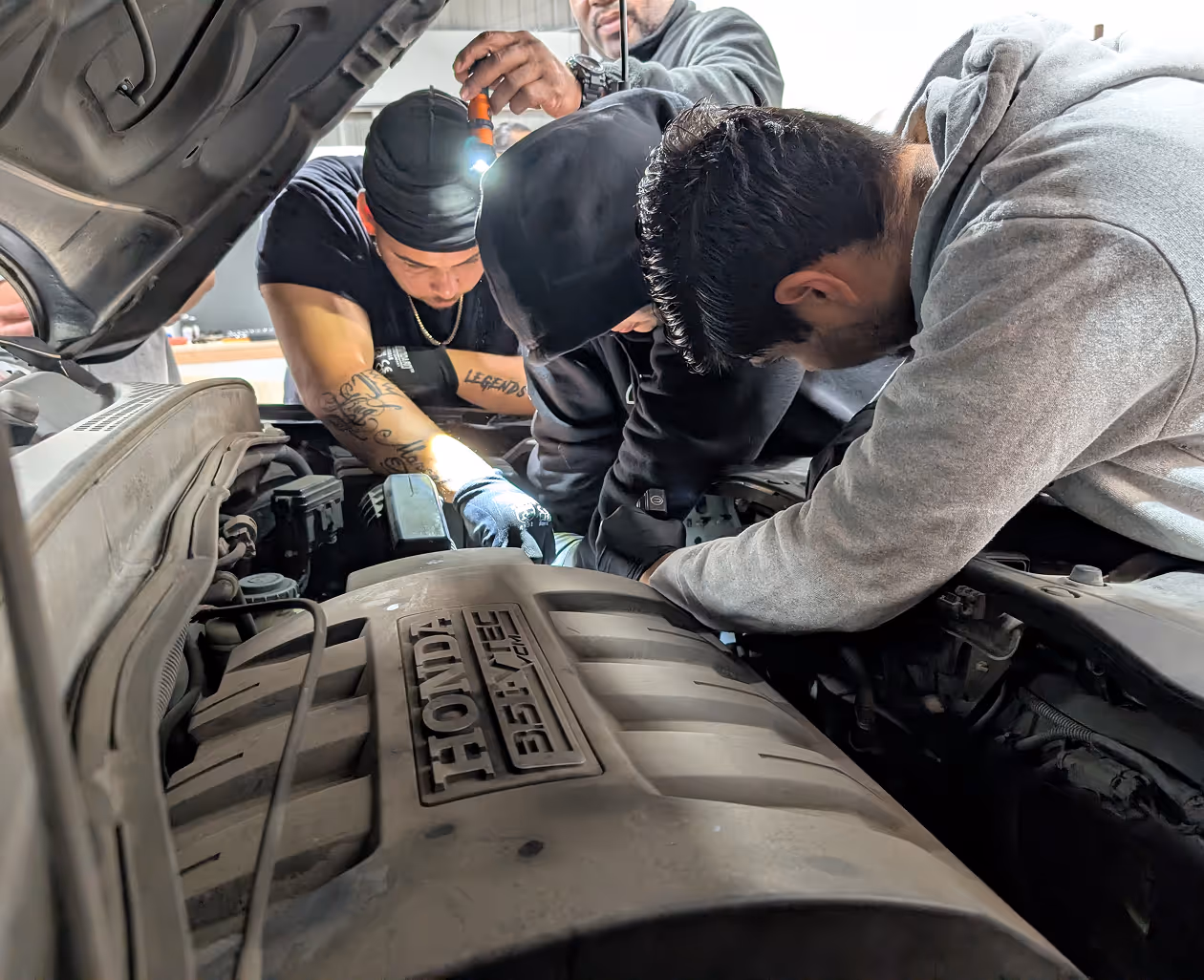 Three men working together under the hood of a Honda vehicle, one holding a flashlight.