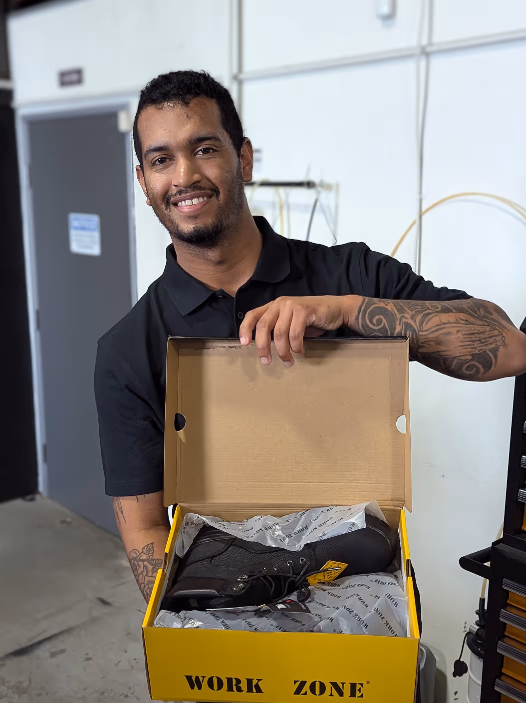 Smiling man with tattoos holding an open yellow Work Zone shoebox displaying a black work boot inside.