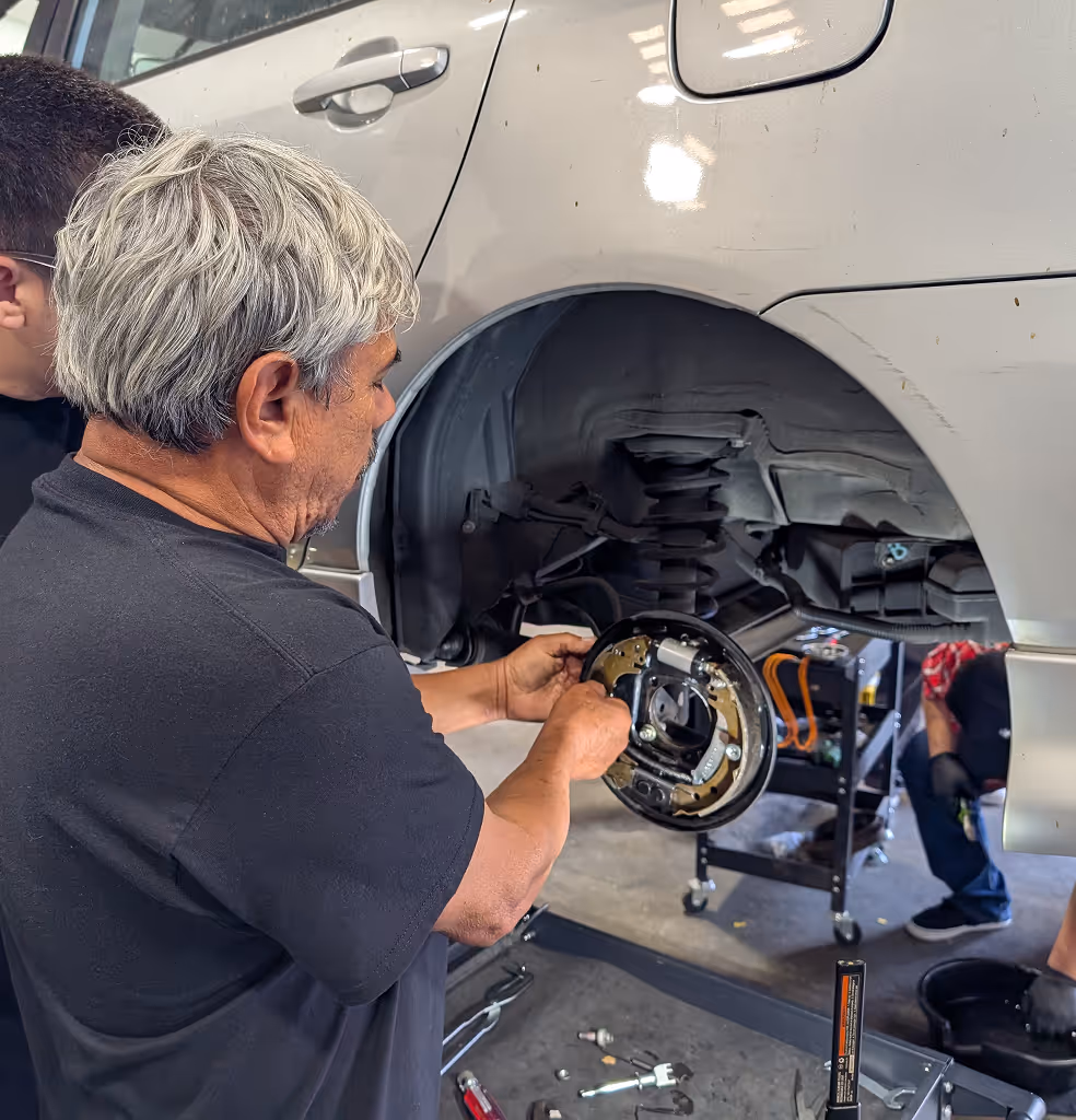 Man working on a car brake assembly with another person assisting nearby in a workshop.