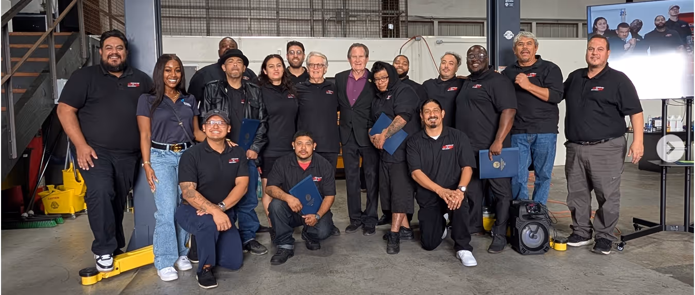 Group of sixteen diverse adults, some holding certificates, posing inside an industrial warehouse with equipment and a screen in the background.
