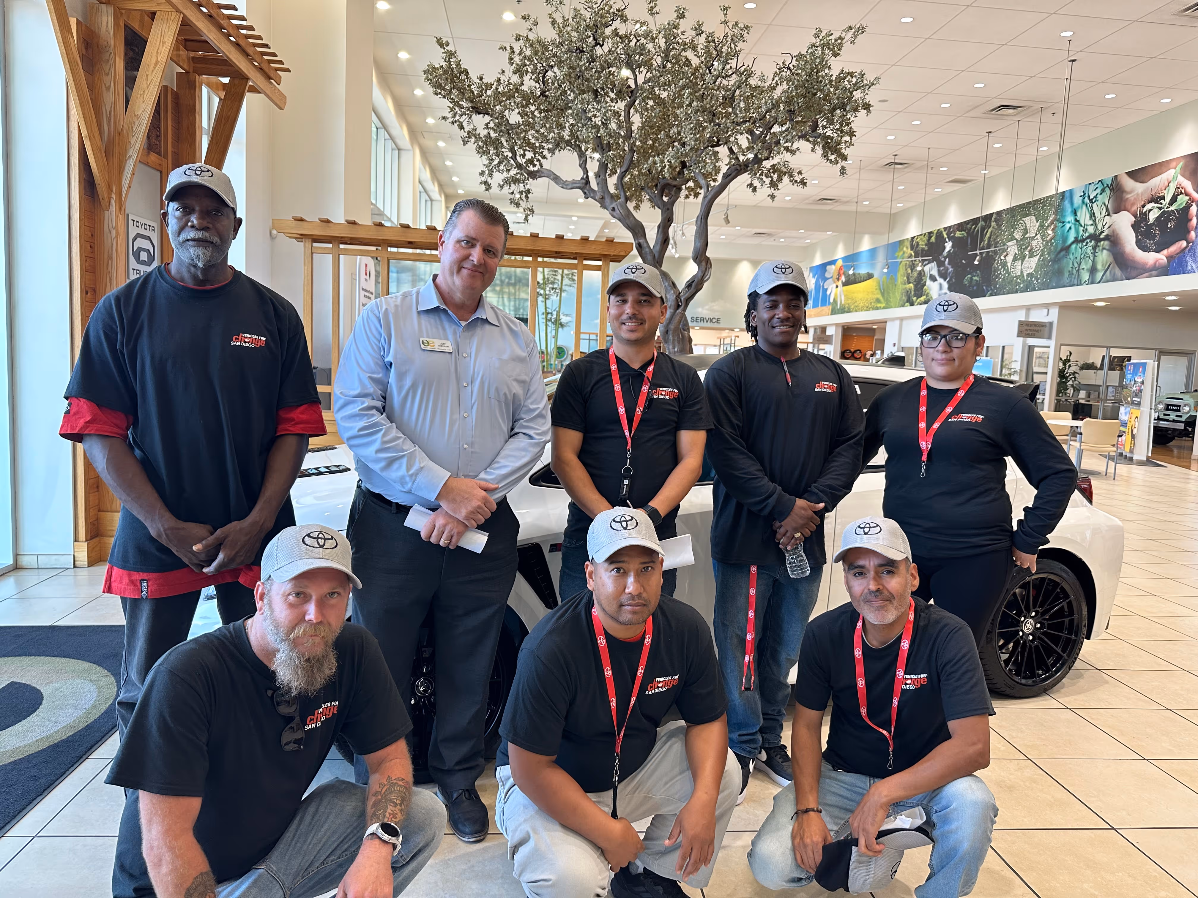 Group of eight people posing inside a Toyota dealership with a white car in the background, most wearing matching black shirts, gray Toyota caps, and red lanyards.