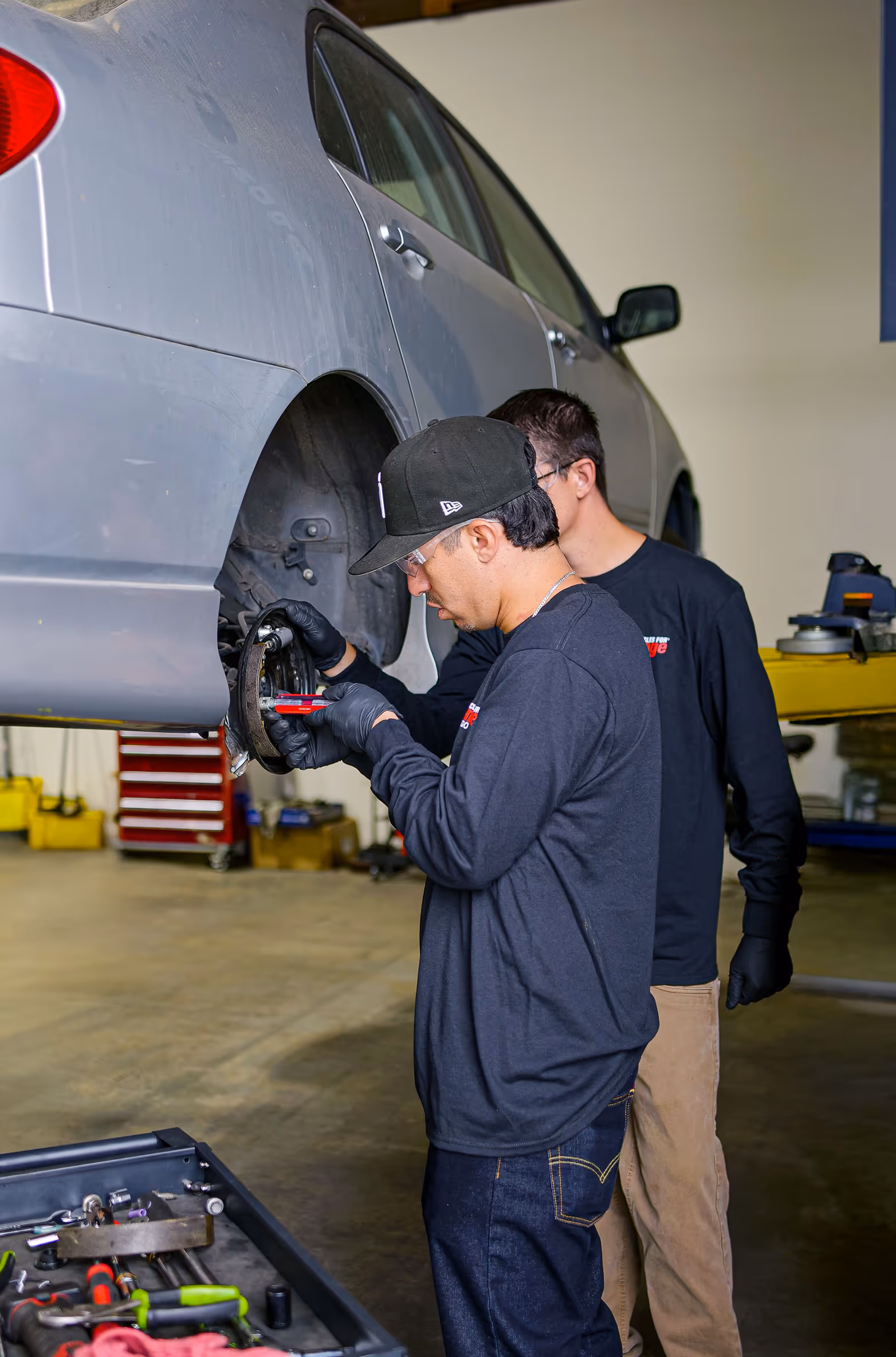 Two mechanics in black shirts and gloves working on the brake assembly of a raised grey car in a workshop.