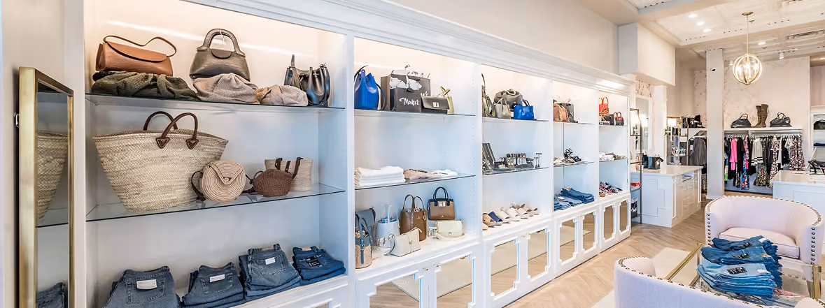 Fashion boutique interior with shelves displaying handbags, folded clothes, shoes, and denim, alongside a seating area and clothing racks.