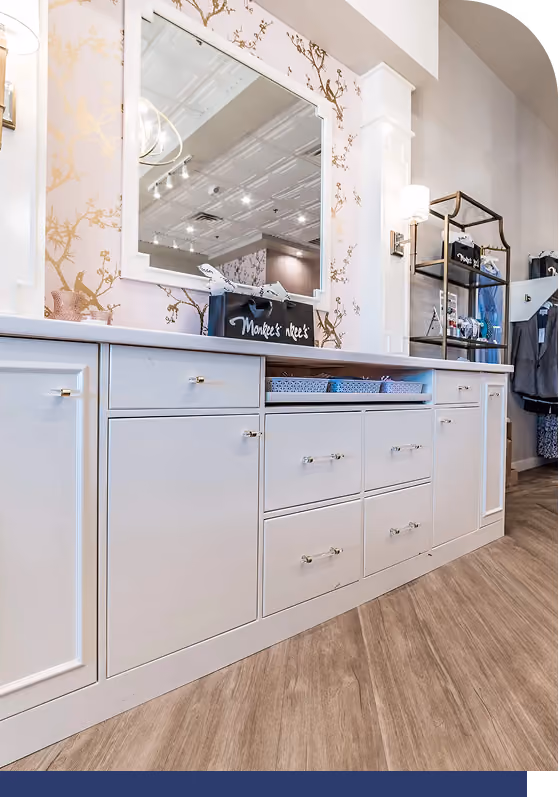 White cabinetry with drawers and shelves below a large mirror mounted on floral wallpaper, featuring a shopping bag labeled 'Monkee's Nook'.