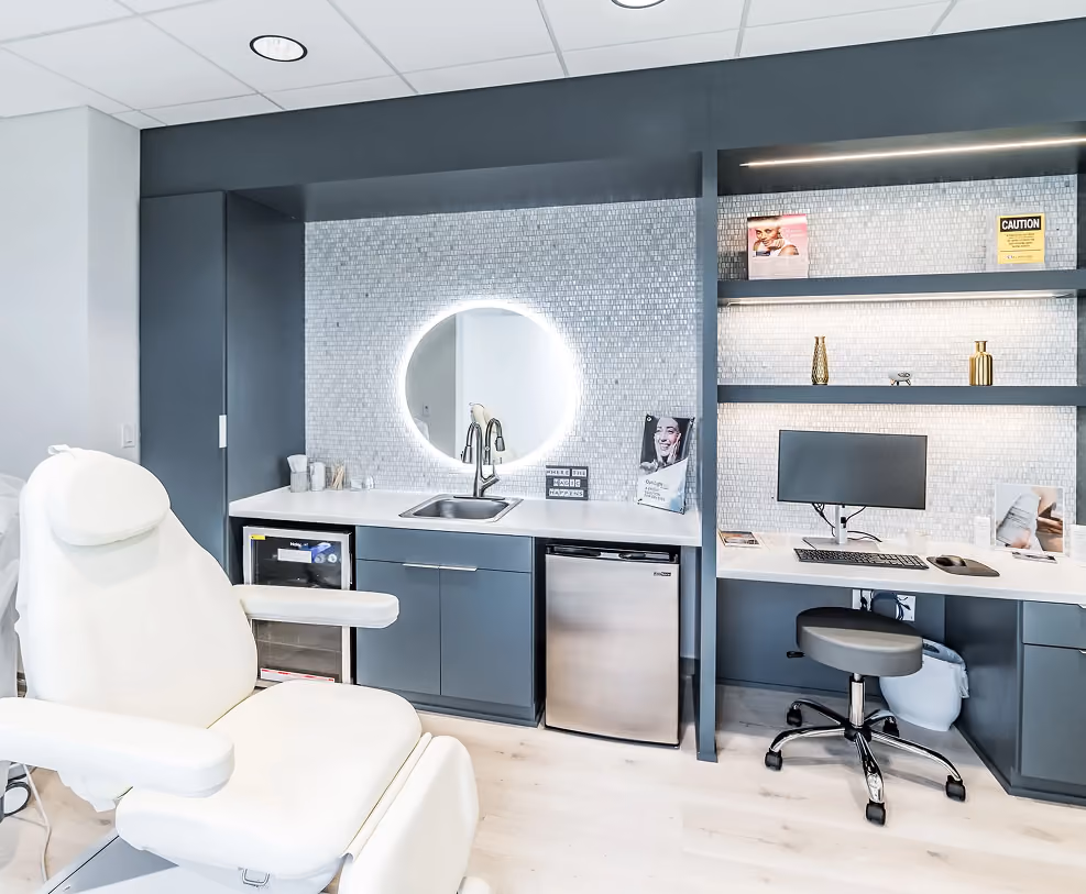 Modern medical or beauty treatment room with white reclining chair, illuminated round mirror, sink, computer desk, and shelves with decorative items.