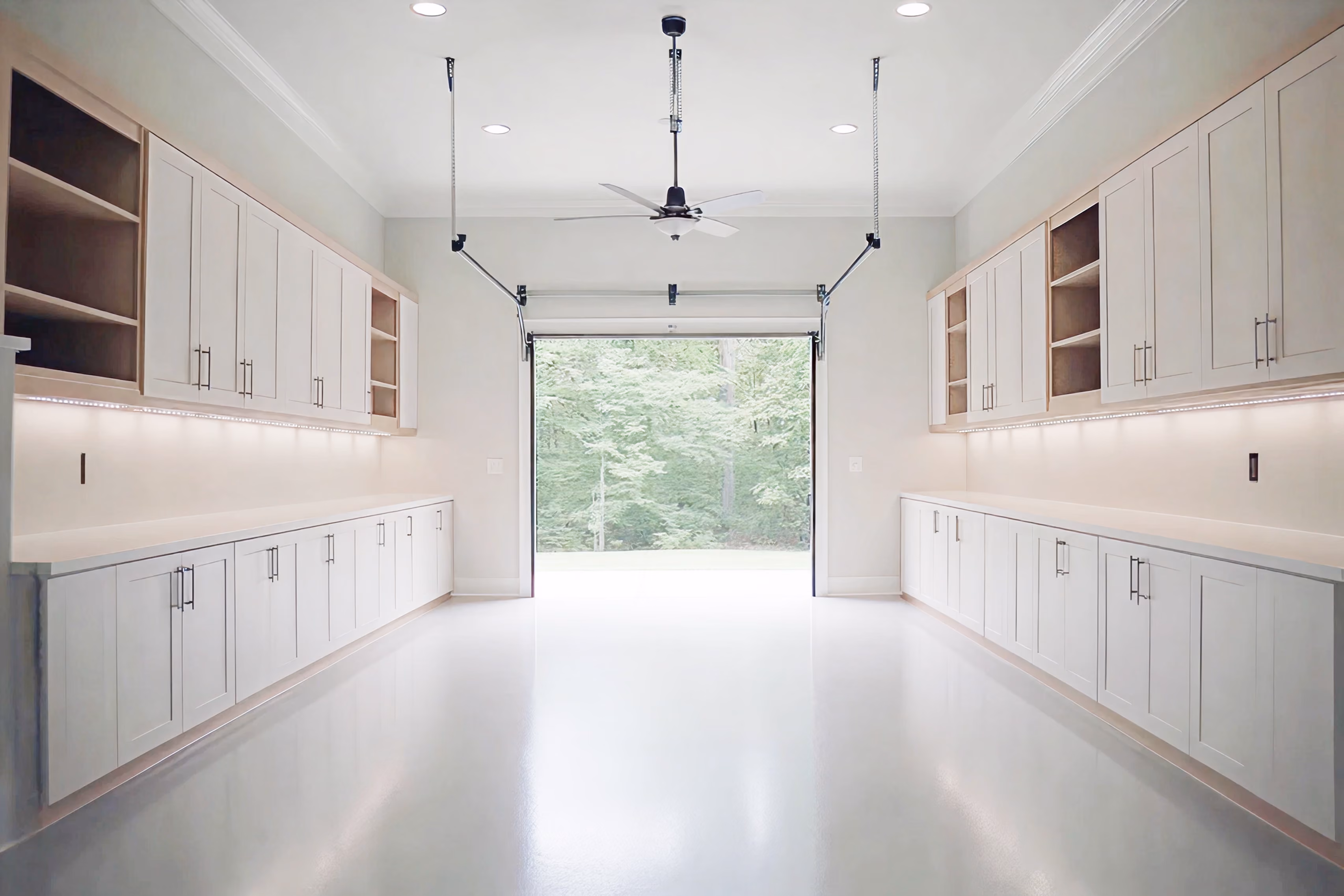 Bright, clean garage with white cabinets lining both walls, a ceiling fan, and an open garage door showing green trees outside.