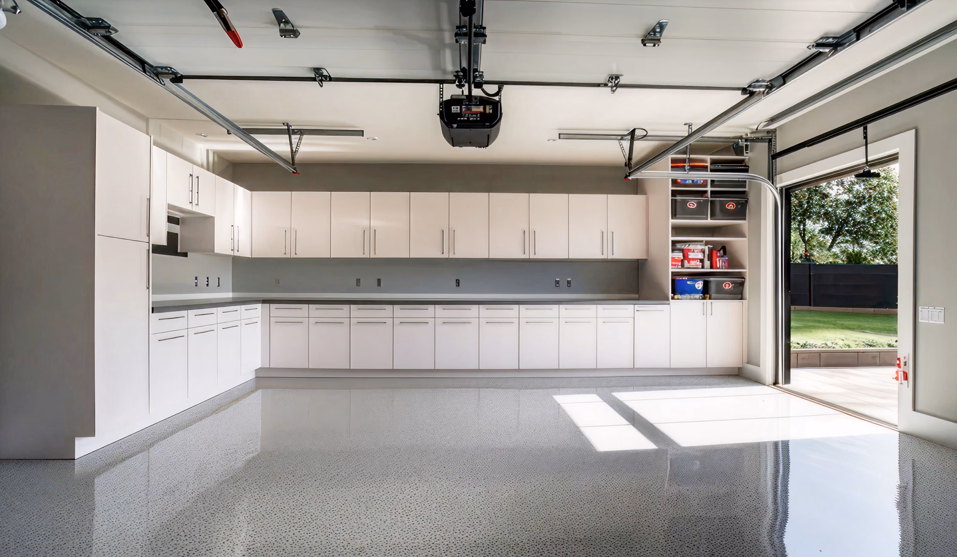 Clean spacious garage with glossy epoxy floor, white cabinets along the walls, and an open garage door showing a green lawn outside.