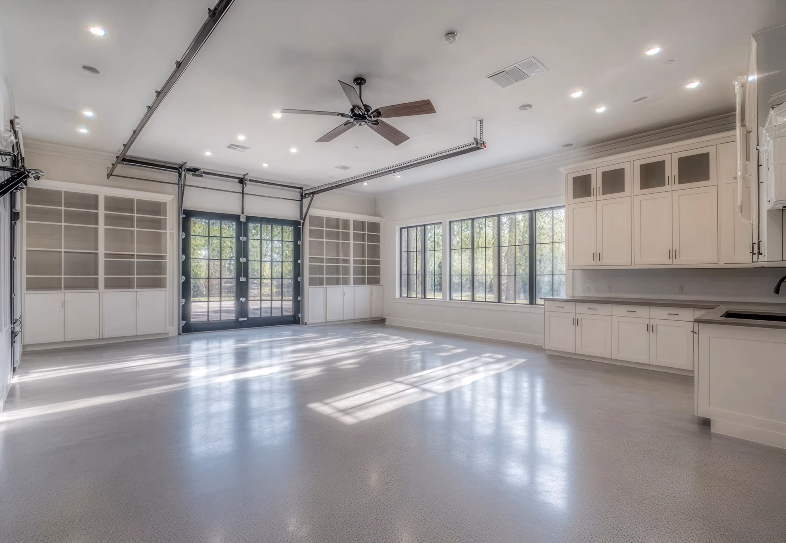 Spacious modern garage with large glass-paneled doors, built-in white cabinets, shelving units, and ceiling fan.