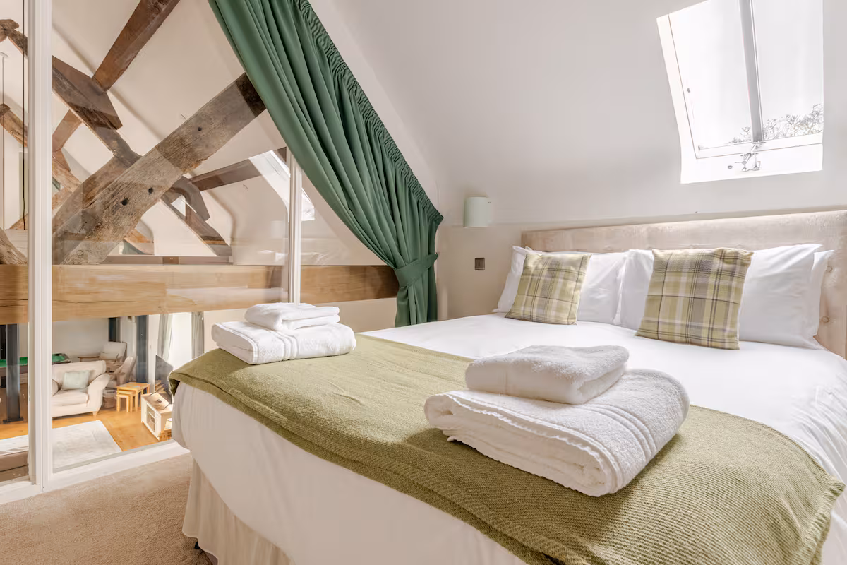 Bright bedroom with a double bed, green throw, plaid pillows, and folded white towels on top, with wooden beams and skylight window.