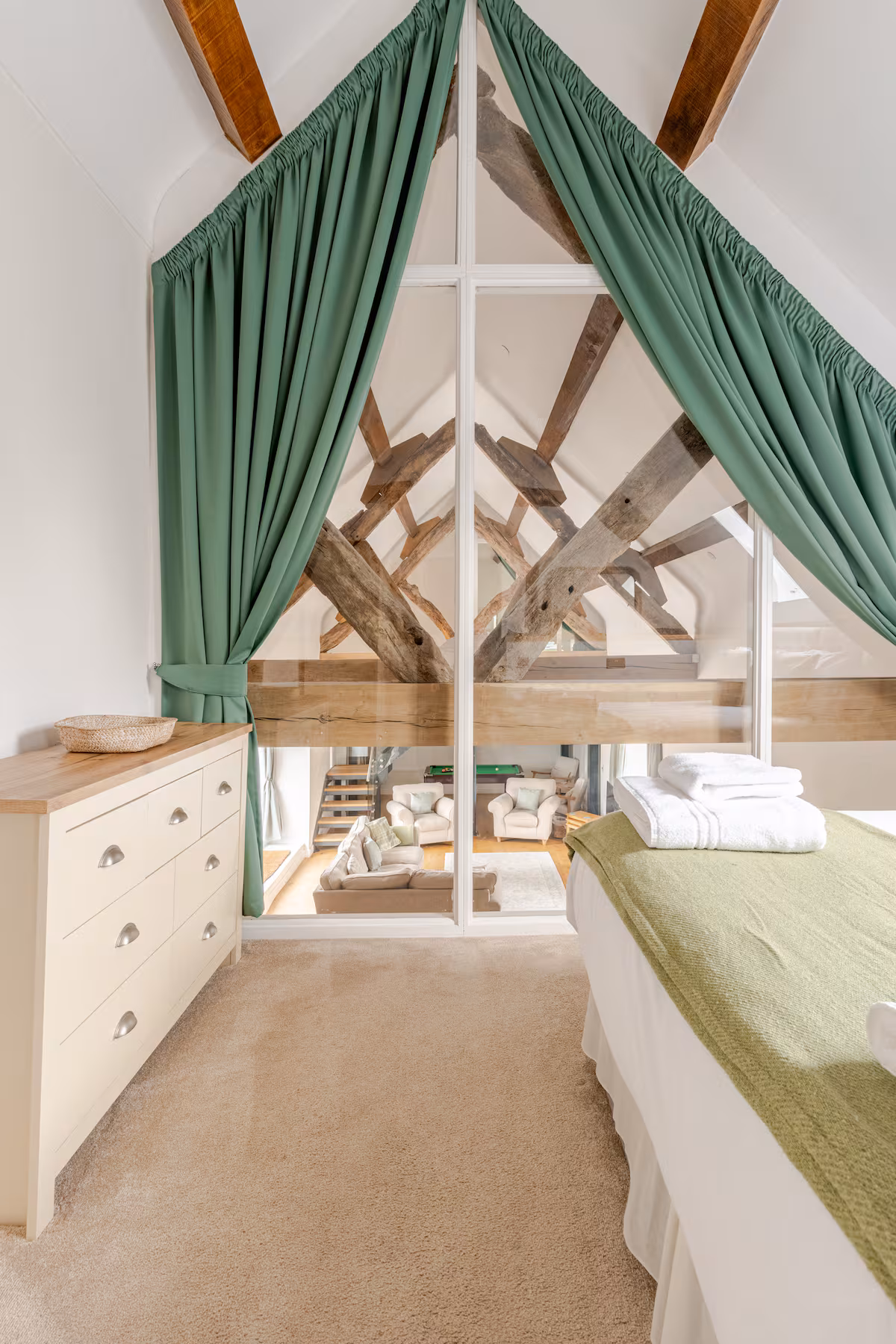Bedroom with green curtains framing a glass partition that shows a living room with exposed wooden beams, beige carpet, white dresser, and a bed with green blanket and folded white towels.
