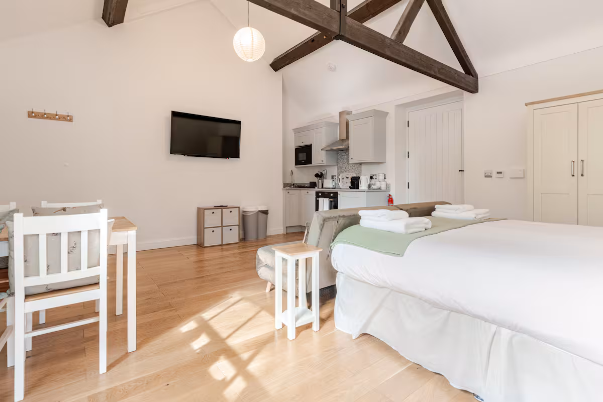 Bright open-plan room with wooden floors, a white bed with folded towels, a small dining table with chairs, a sofa, wall-mounted TV, and kitchenette under exposed wooden beams.