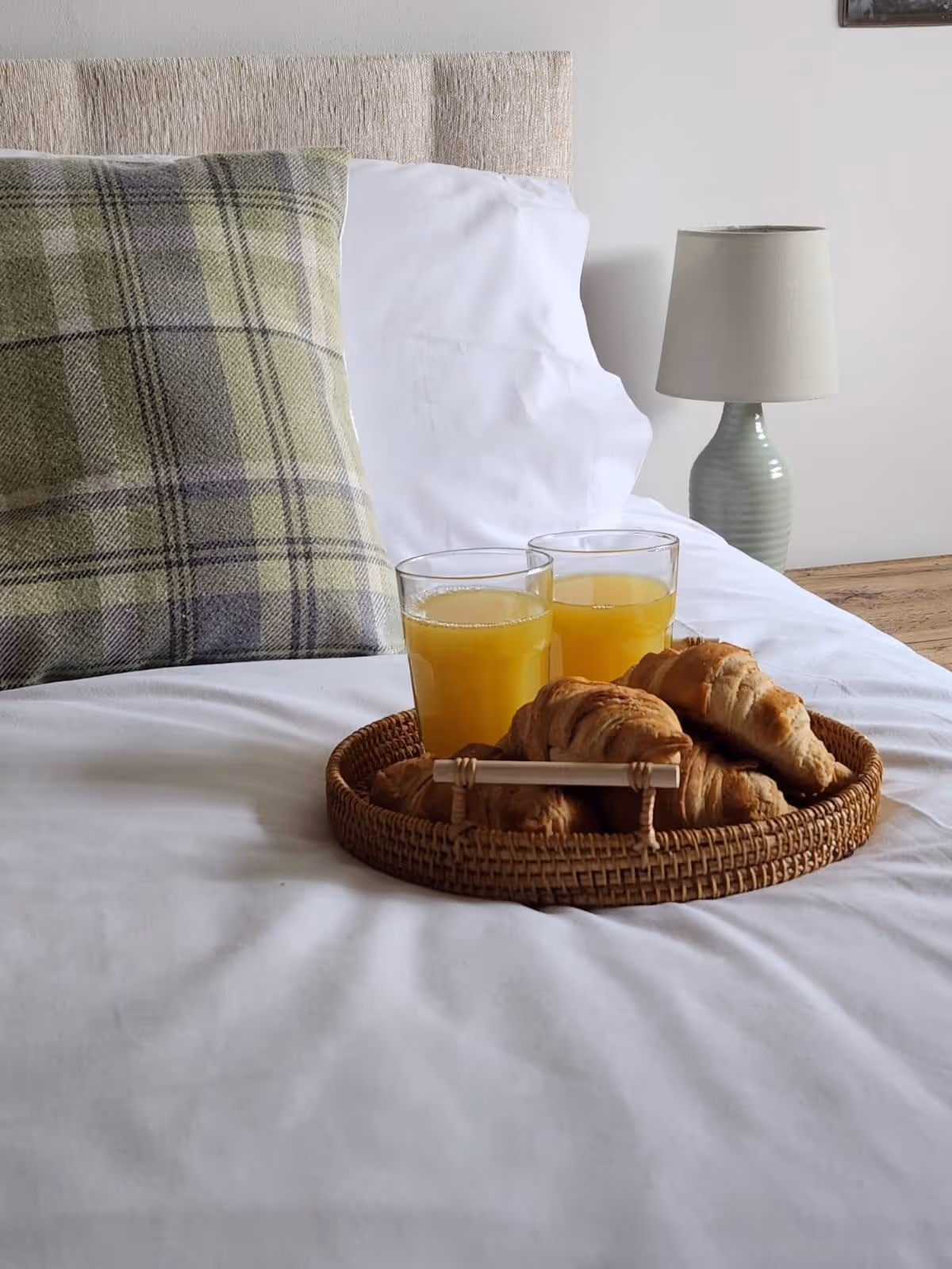 Wicker tray with croissants and two glasses of orange juice on a bed with a white sheet and a plaid pillow, next to a bedside lamp.