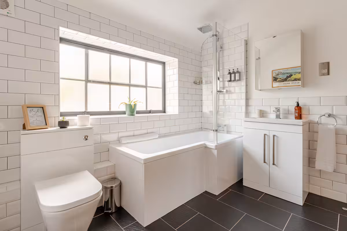 Modern bathroom with white subway tiled walls, an L-shaped bathtub with glass shower screen, a white vanity with mirror, and a toilet under a large frosted window.