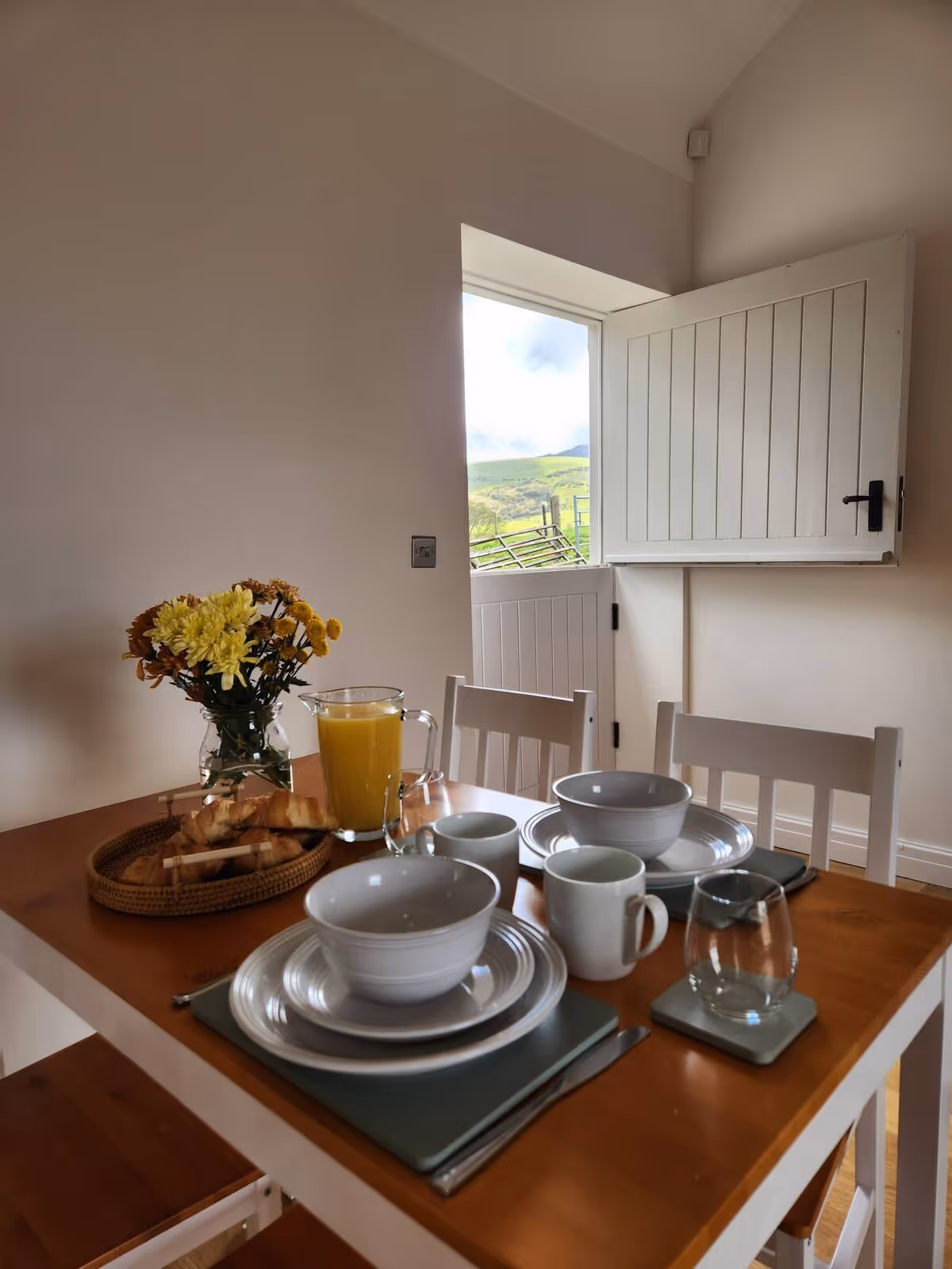 Wooden dining table set for two with white dishes, mugs, a glass of orange juice, a basket of croissants, and a vase of yellow flowers under a window with a countryside view.