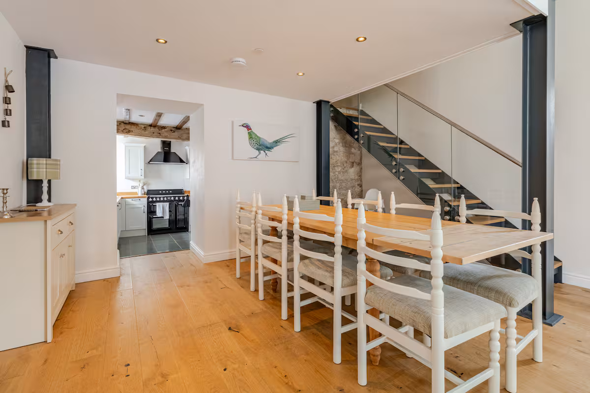 Dining area with wooden table, white cushioned chairs, staircase with glass railing, and view into kitchen with black stove.
