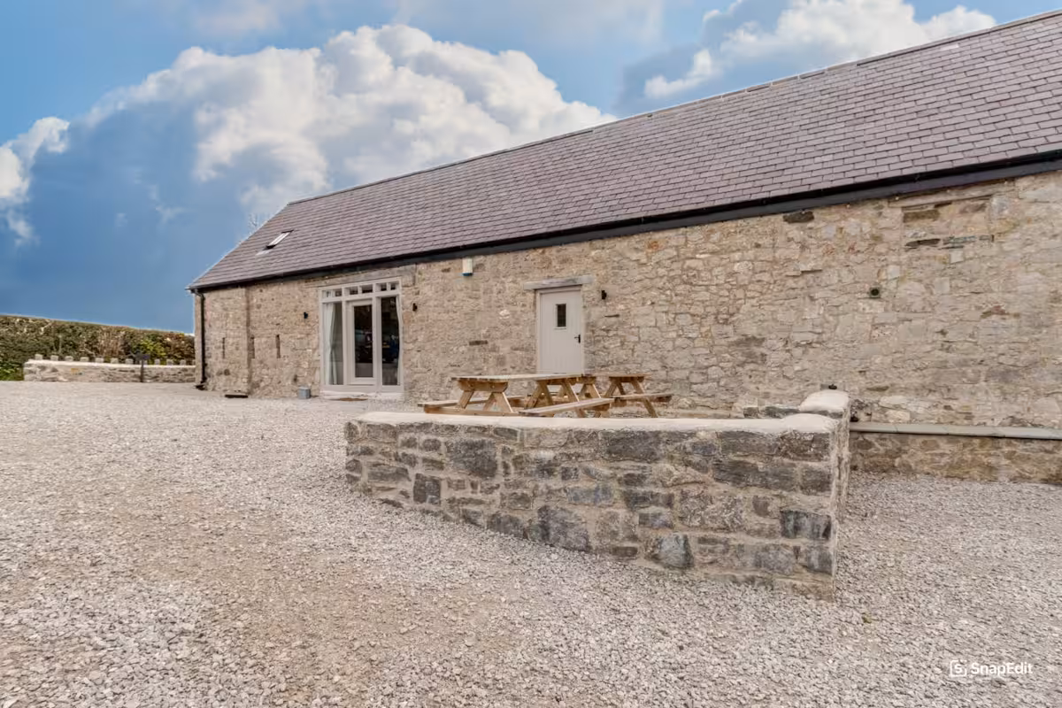 Long stone building with slate roof, gravel courtyard, picnic tables, and cloudy blue sky.