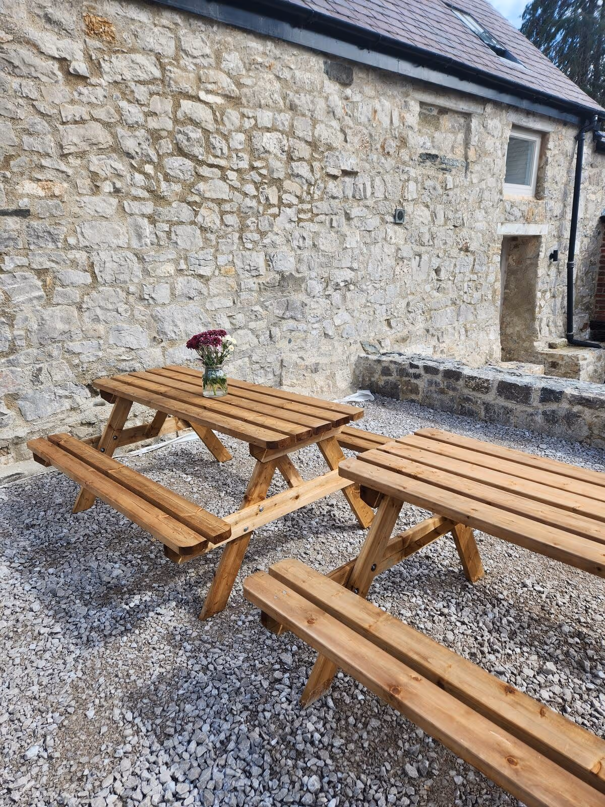 Two wooden picnic tables on gravel beside a stone building, one with a small vase of flowers on top.