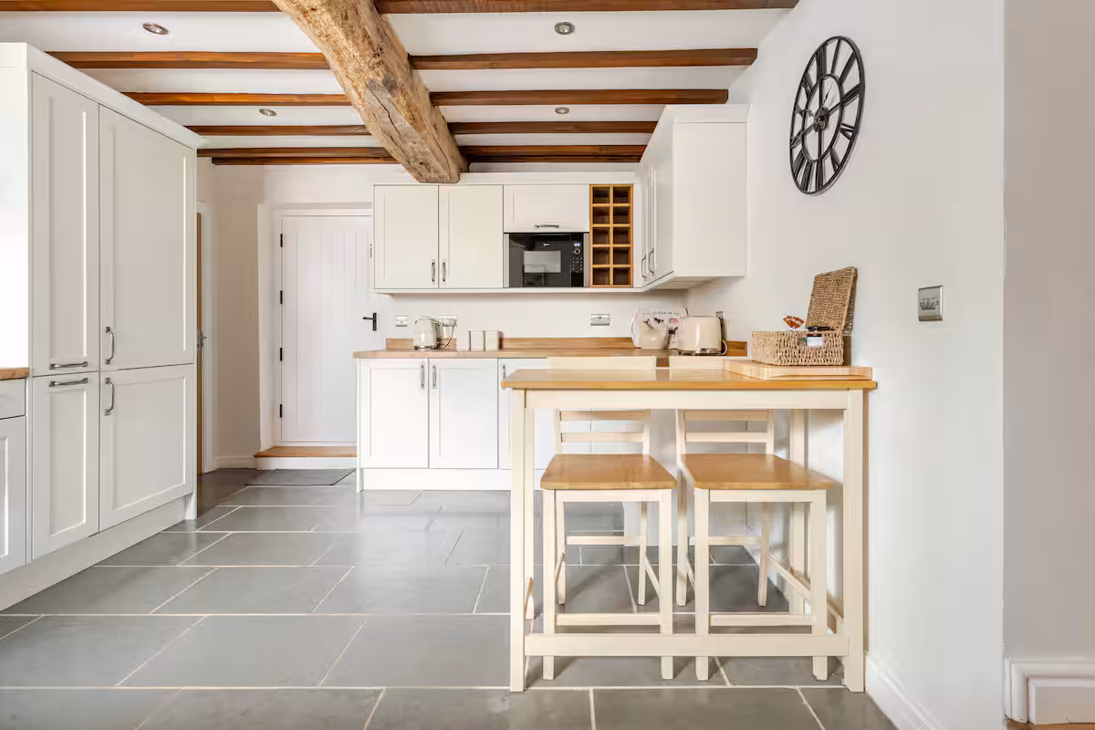 Bright kitchen with white cabinets, wooden countertops, exposed wooden ceiling beams, a wall clock, and a small wooden table with two chairs.