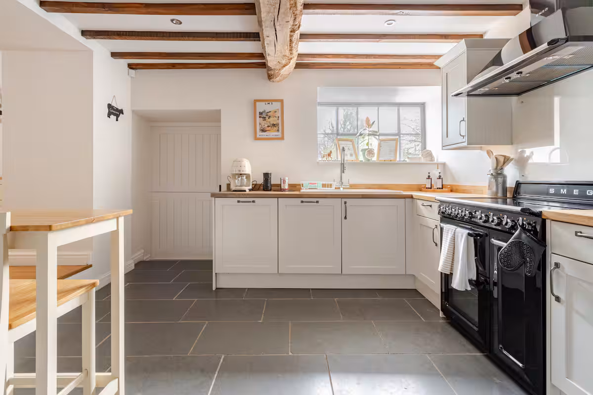 Bright kitchen with wooden beams, white cabinets, black SMEG stove, and a sink under a window with decorative items.