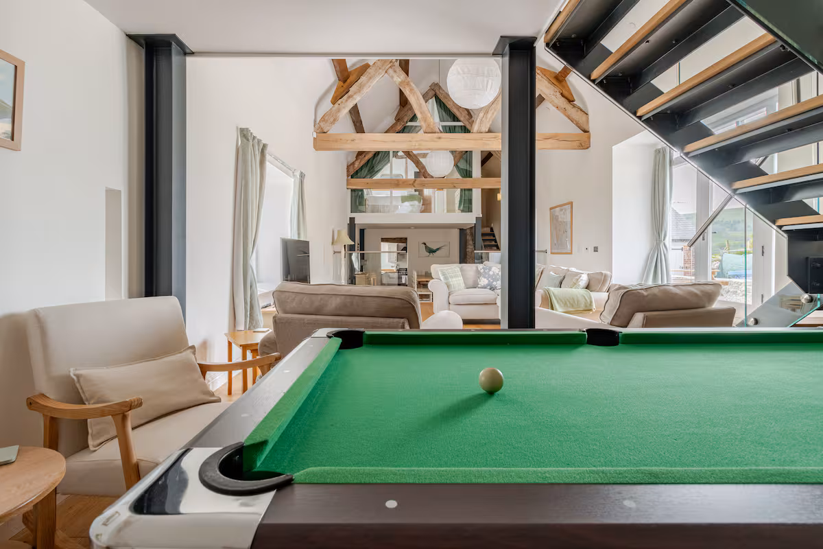 Interior of a modern living room with exposed wooden beams, beige sofas, and a pool table with a green felt surface and a single white ball in the center.