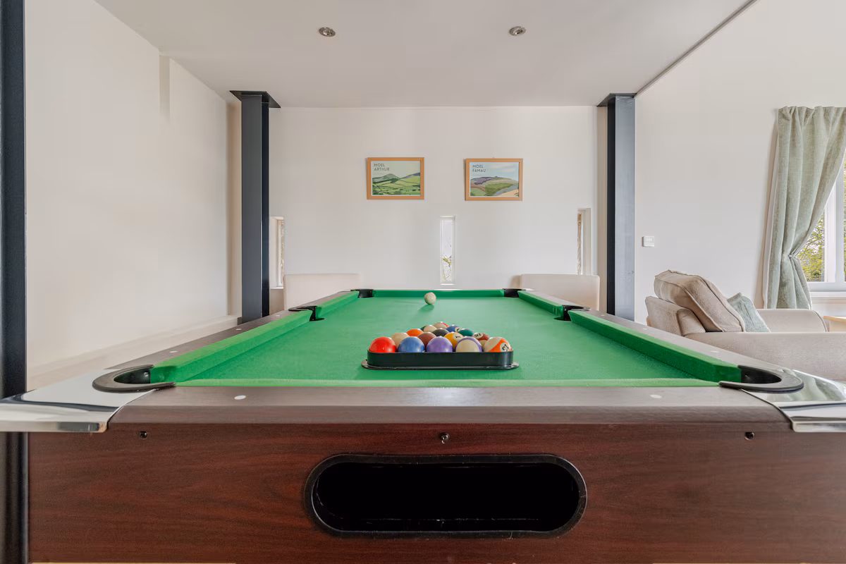 View down a pool table with racked colorful billiard balls in a bright, modern room with white walls, two framed pictures, and beige furniture.