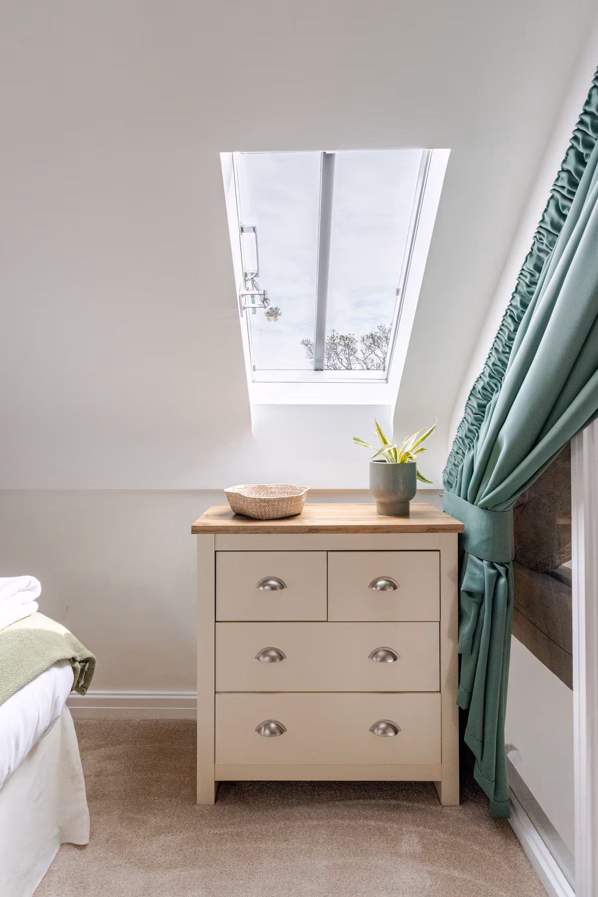 Light beige chest of drawers under a skylight window with a woven basket and a potted plant on top, next to a bed with green blanket and green curtains.