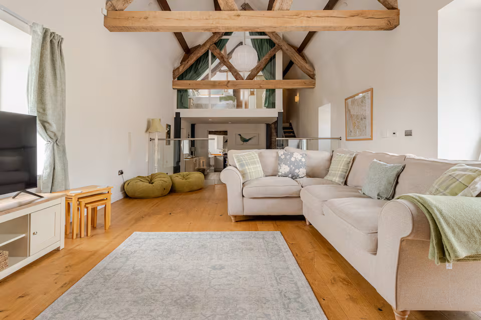 Bright lounge room with beige sectional sofa, wooden beams, light hardwood floor, TV, and green accents.
