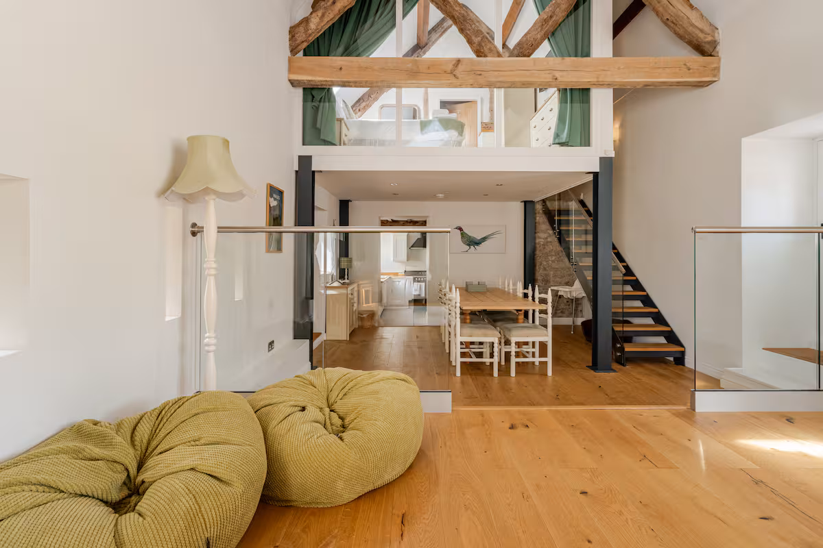 Modern two-level living area with wooden beams, green bean bag chairs, dining table with white chairs, and staircase to upper bedroom.
