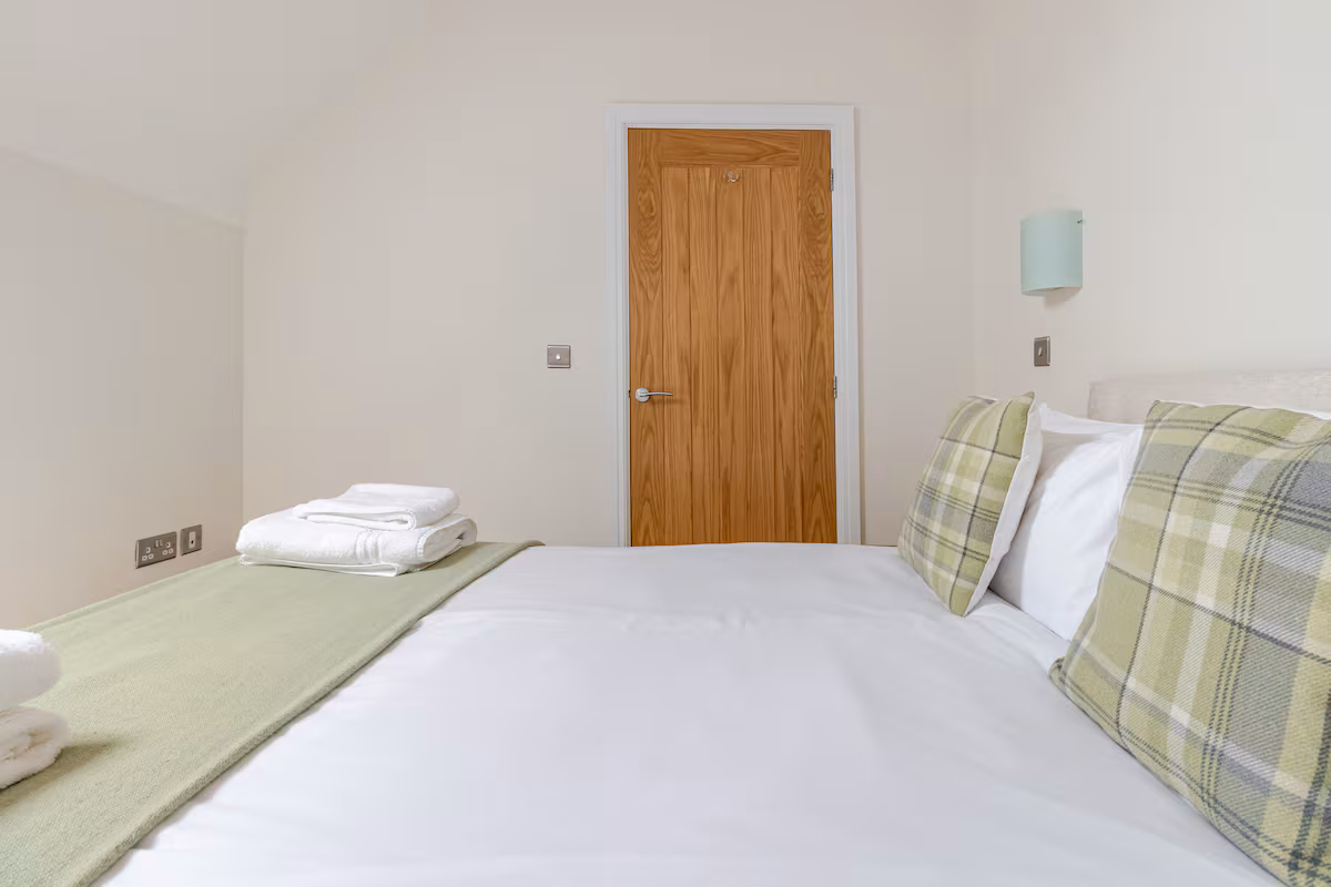 Minimalist bedroom with a neatly made bed featuring white linens, green plaid pillows, and folded towels, facing a wooden door.