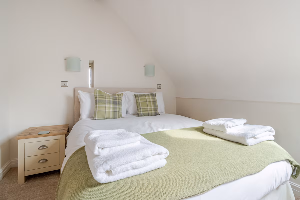 Bedroom with a double bed topped with white linens, green plaid pillows, green throw blanket, and folded white towels on the foot of the bed.