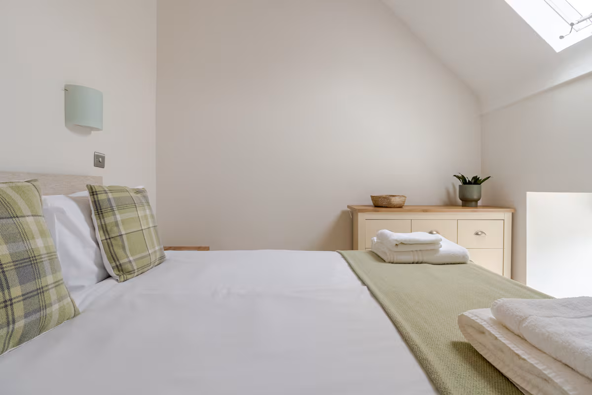 Minimalist bedroom with white bed linens, green plaid pillows, folded towels, and a wooden dresser with a plant and basket under a skylight.
