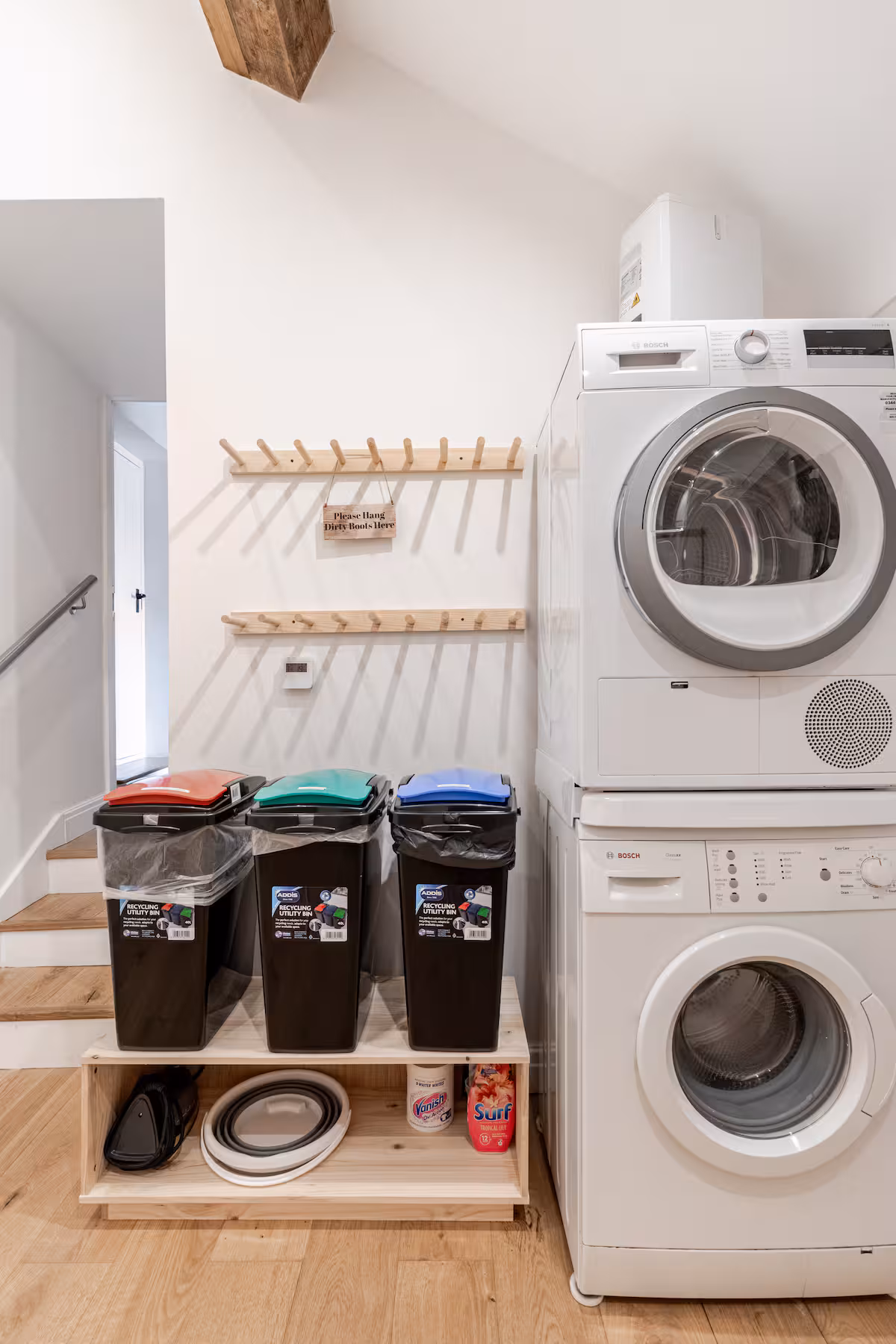 Laundry room with stacked Bosch washer and dryer next to three recycling bins with colored lids on a wooden shelf, and a wall-mounted rack with a sign reading 'Please Hang Dirty Boots Here'.
