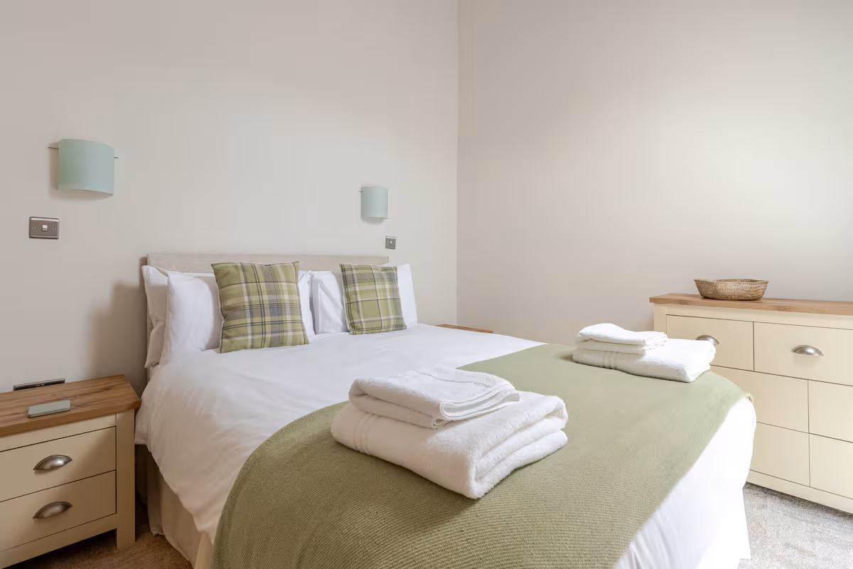 Neatly made bed with white linens, green plaid pillows, and folded white towels on a green blanket in a minimalist bedroom.
