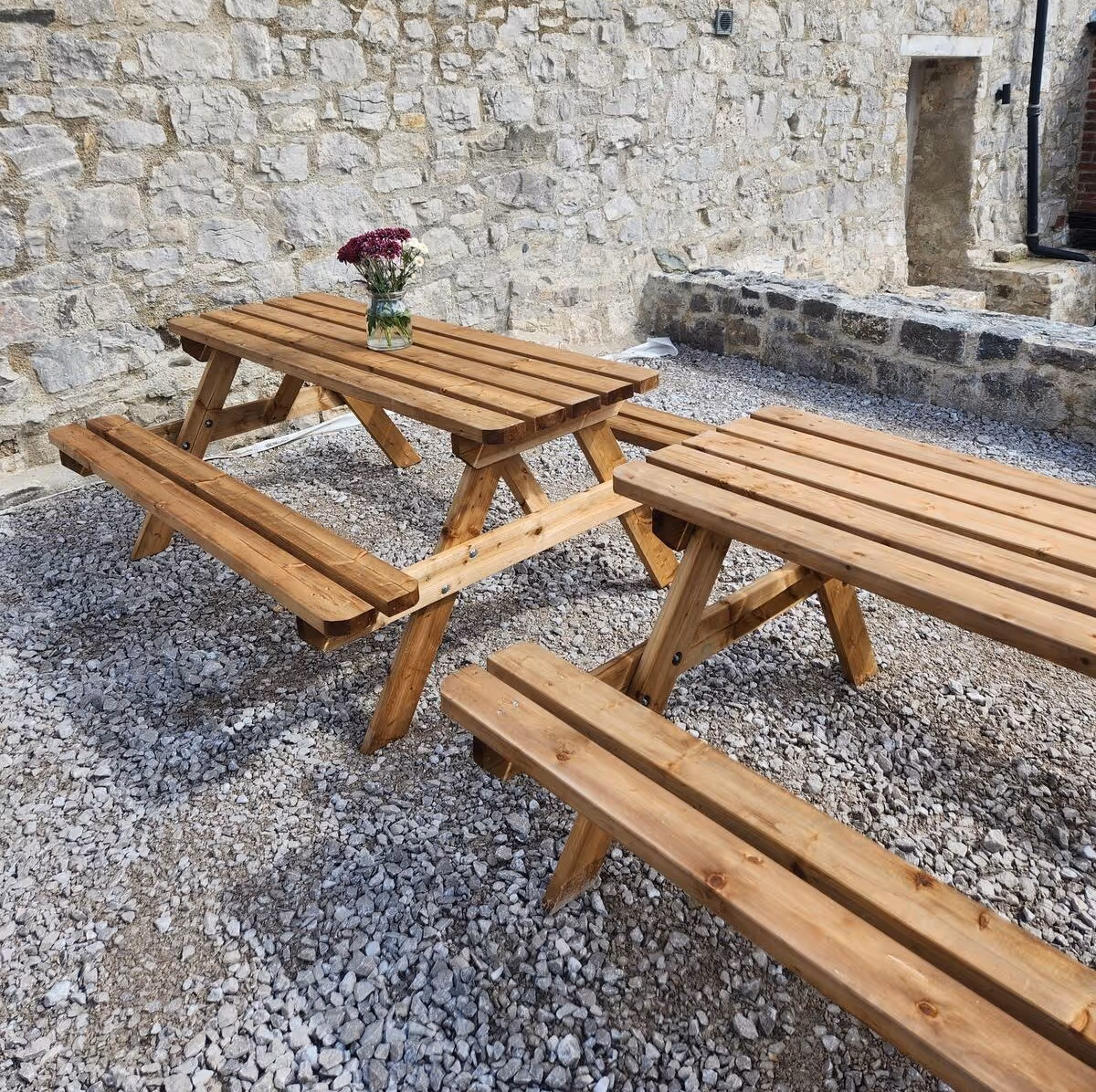 Two wooden picnic tables with attached benches on gravel ground, one table has a small vase with flowers on it, against a stone wall background.