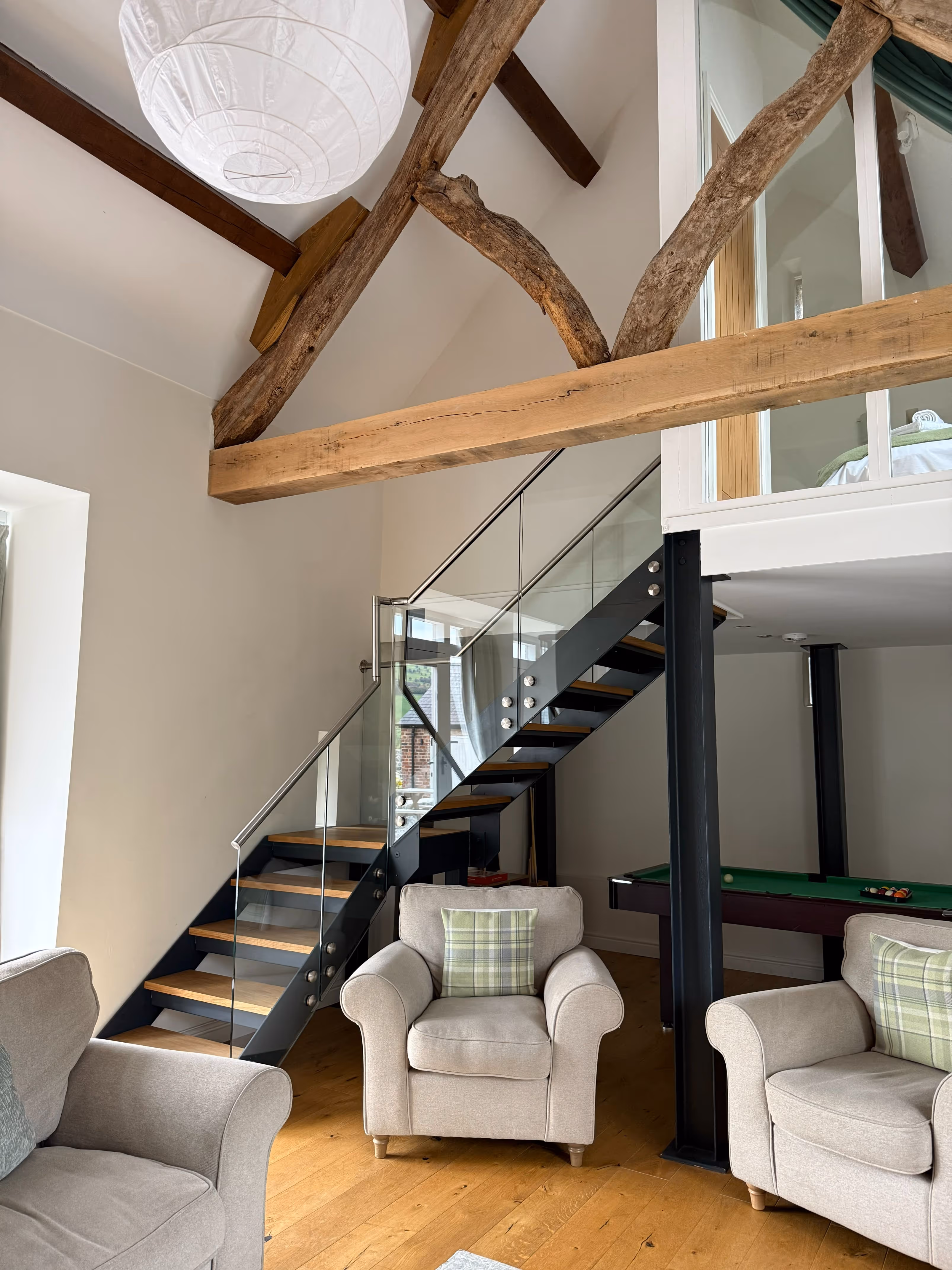 Modern living room with beige armchairs featuring green plaid pillows, wooden floor, exposed wooden ceiling beams, a glass and metal staircase, and a pool table under a loft area.