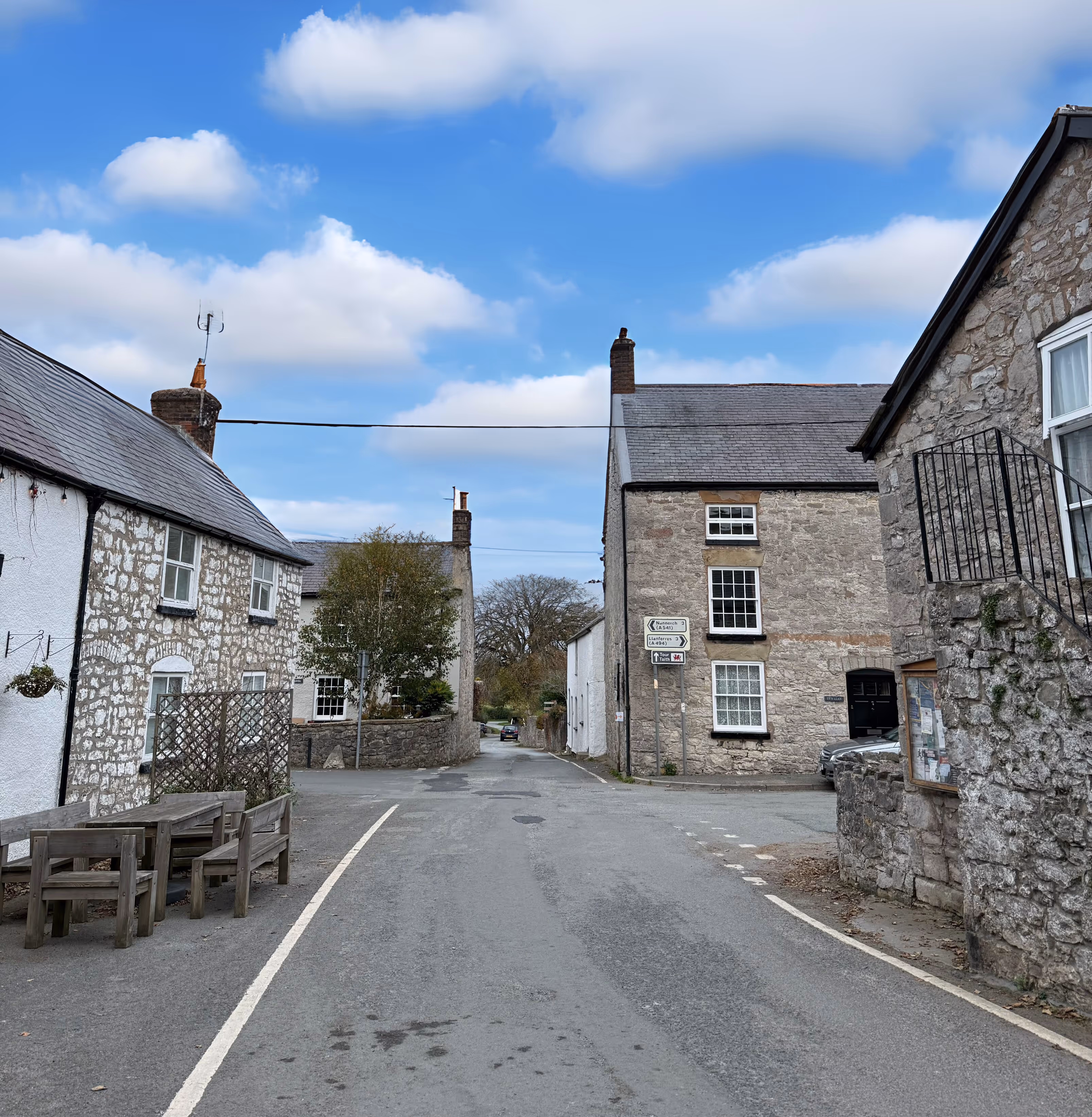 Quiet village street lined with stone and whitewashed houses under a partly cloudy blue sky.