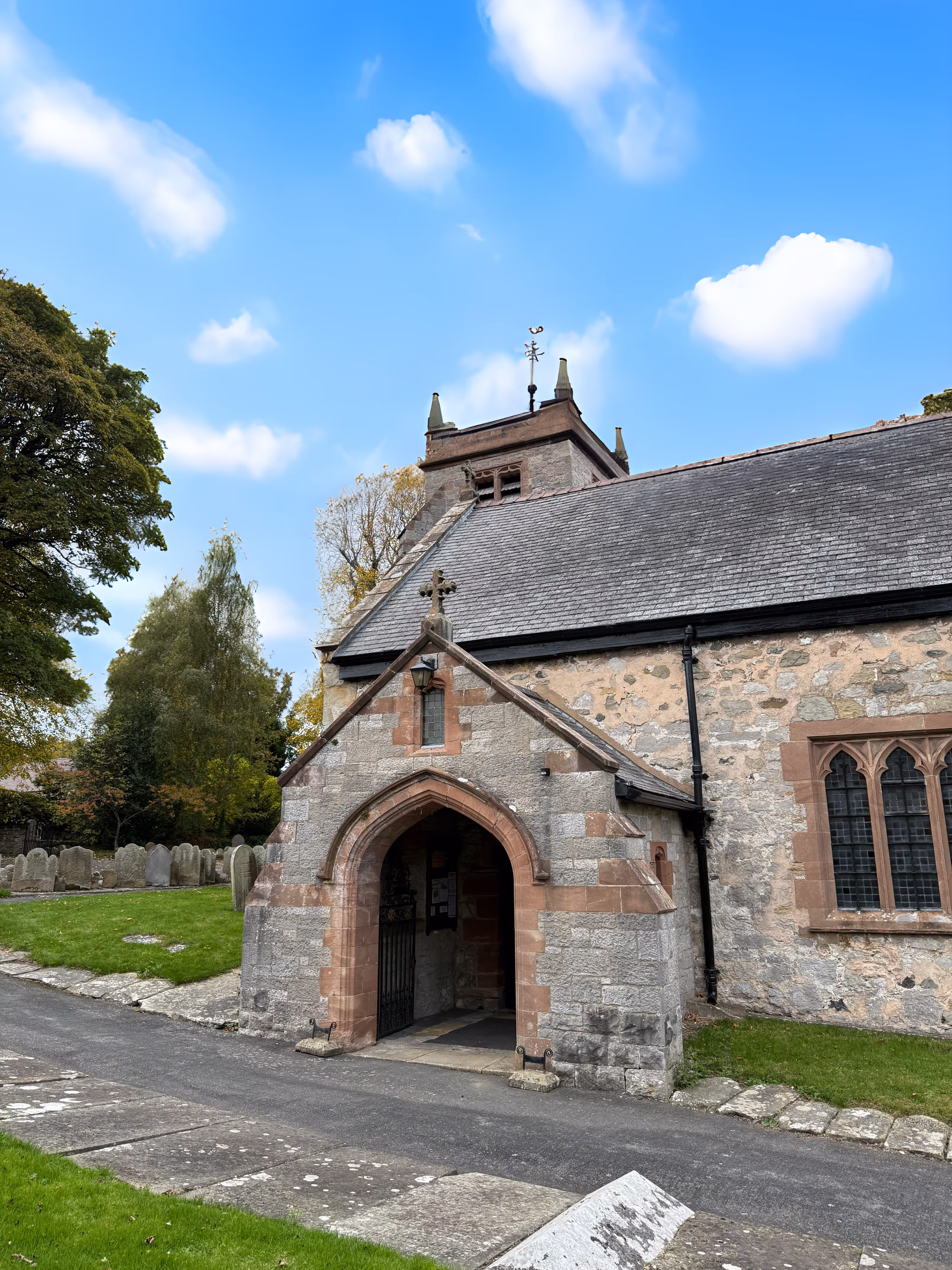 Stone church entrance with arched doorway and cross above, surrounded by green grass and gravestones under a blue sky.