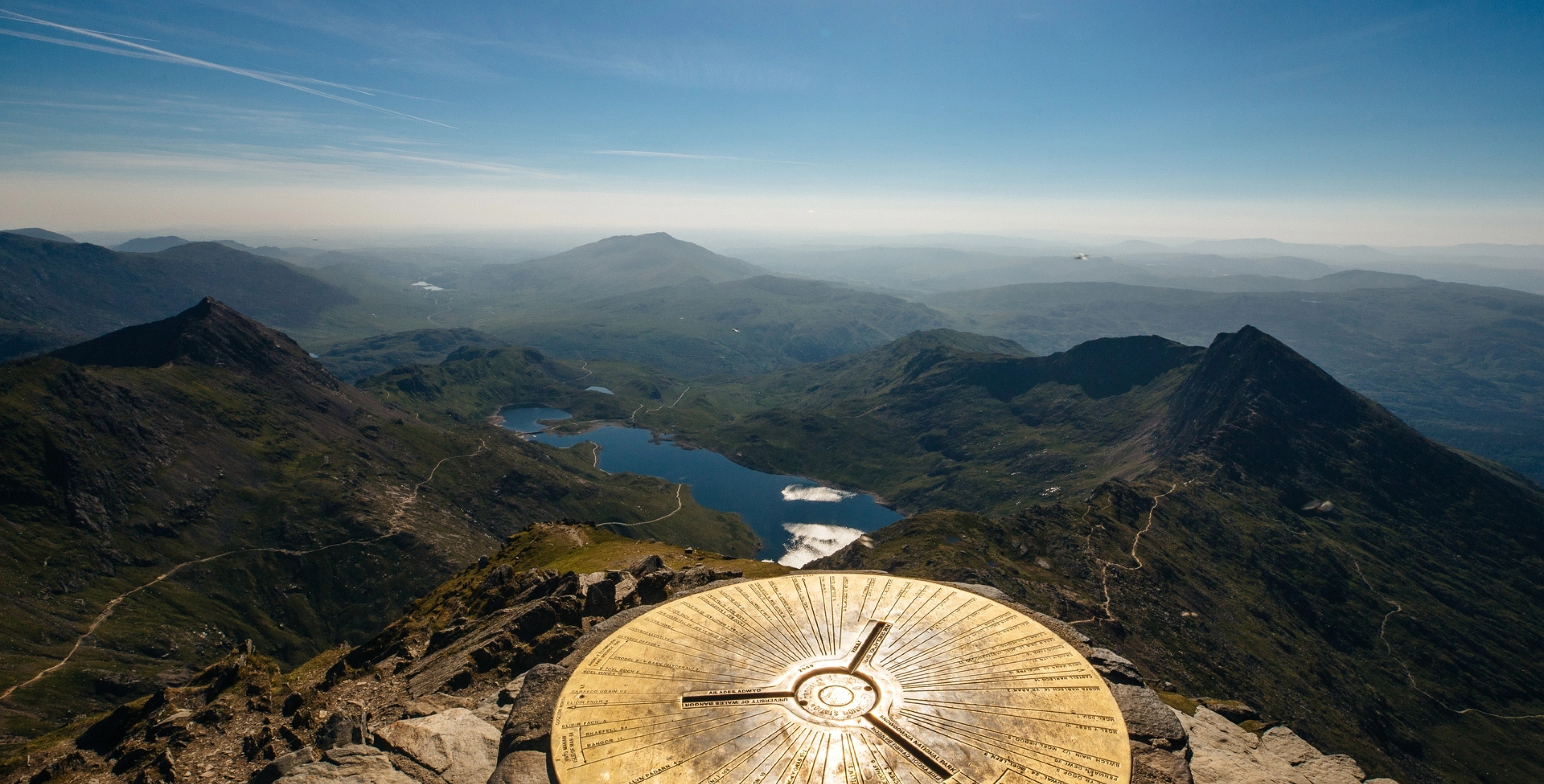 Panoramic view of mountainous landscape with a lake, seen from a summit with a circular brass viewpoint indicator.