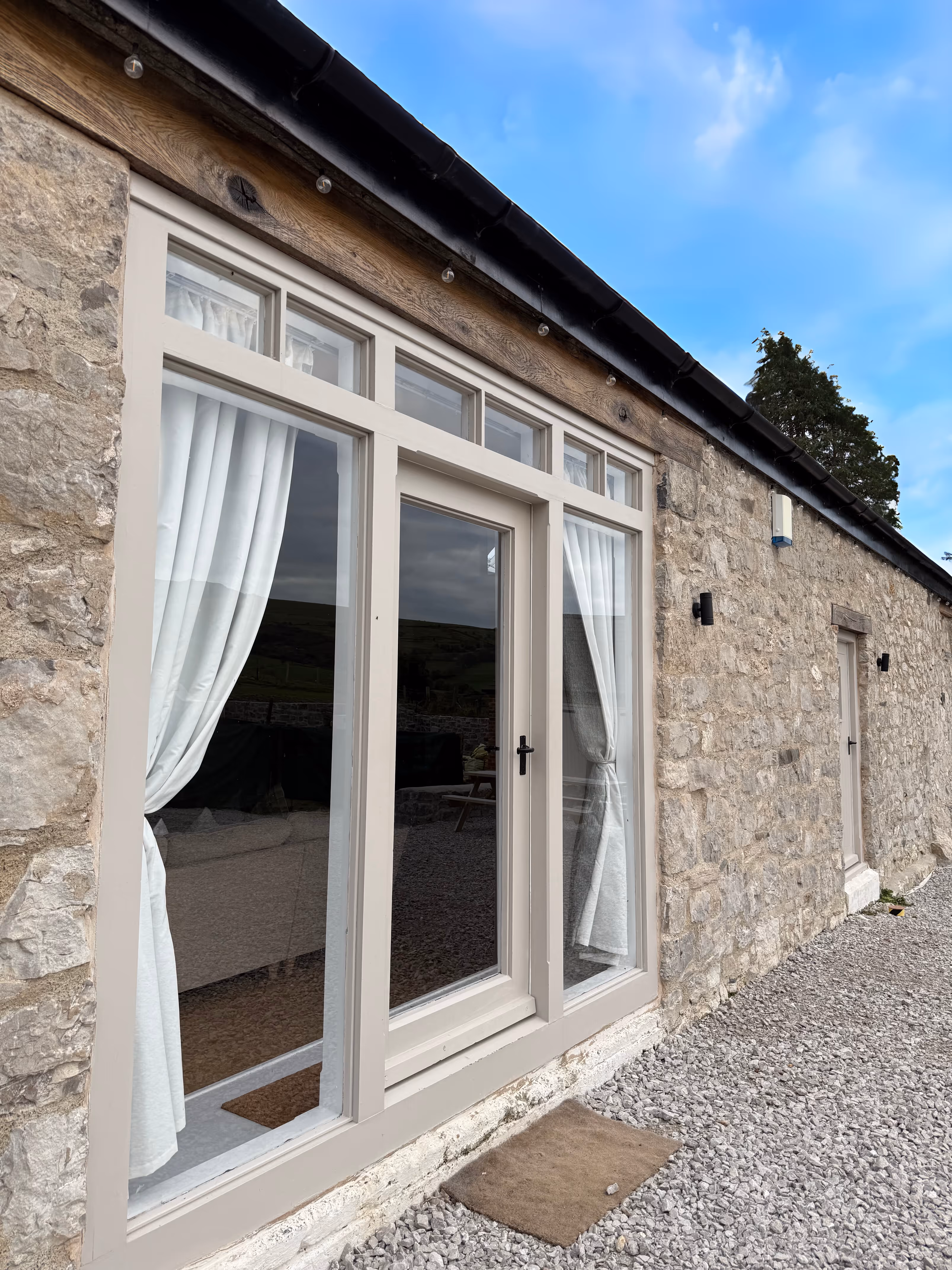 Stone cottage exterior with beige-framed glass door and windows with white curtains, gravel pathway, and blue sky.