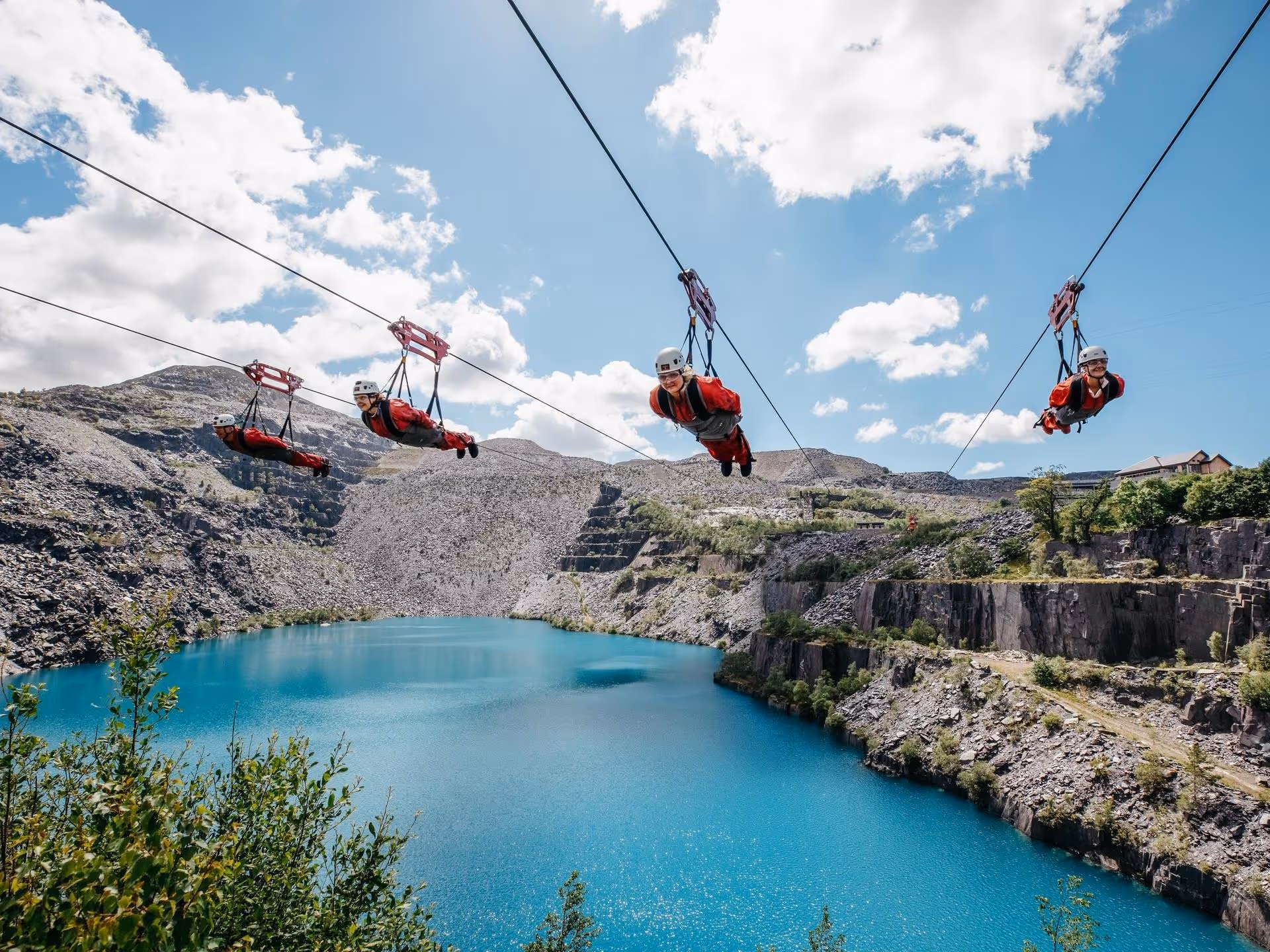 Four people zip lining over a bright blue lake surrounded by rocky cliffs and greenery under a partly cloudy sky.