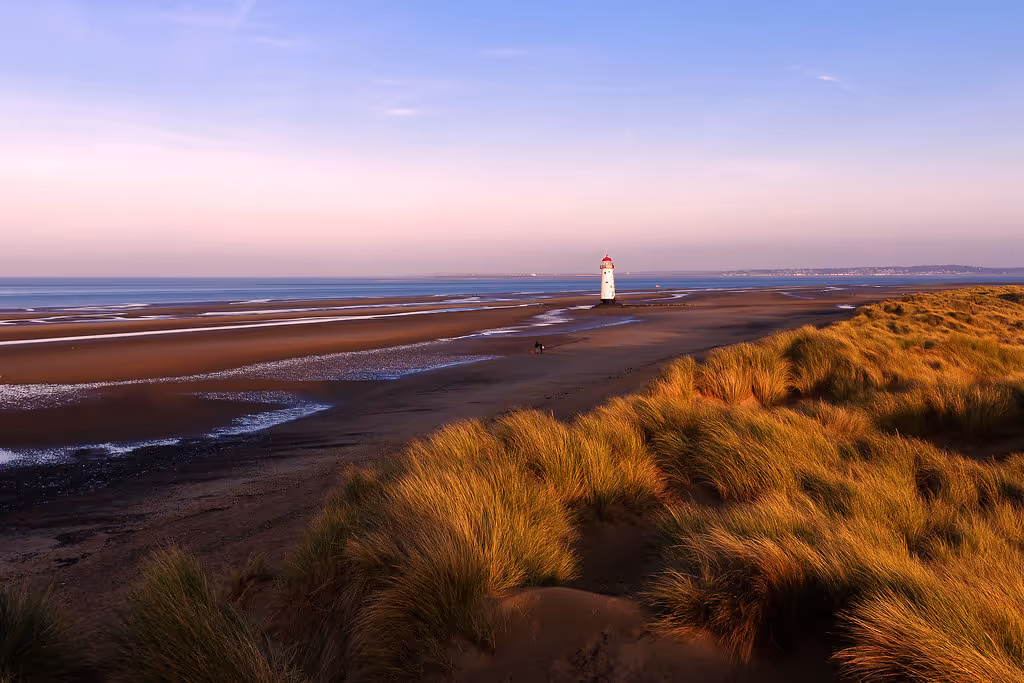 Coastal scene with tall grass dunes in foreground, a sandy beach, and a white lighthouse with a red top near the shore at sunset.