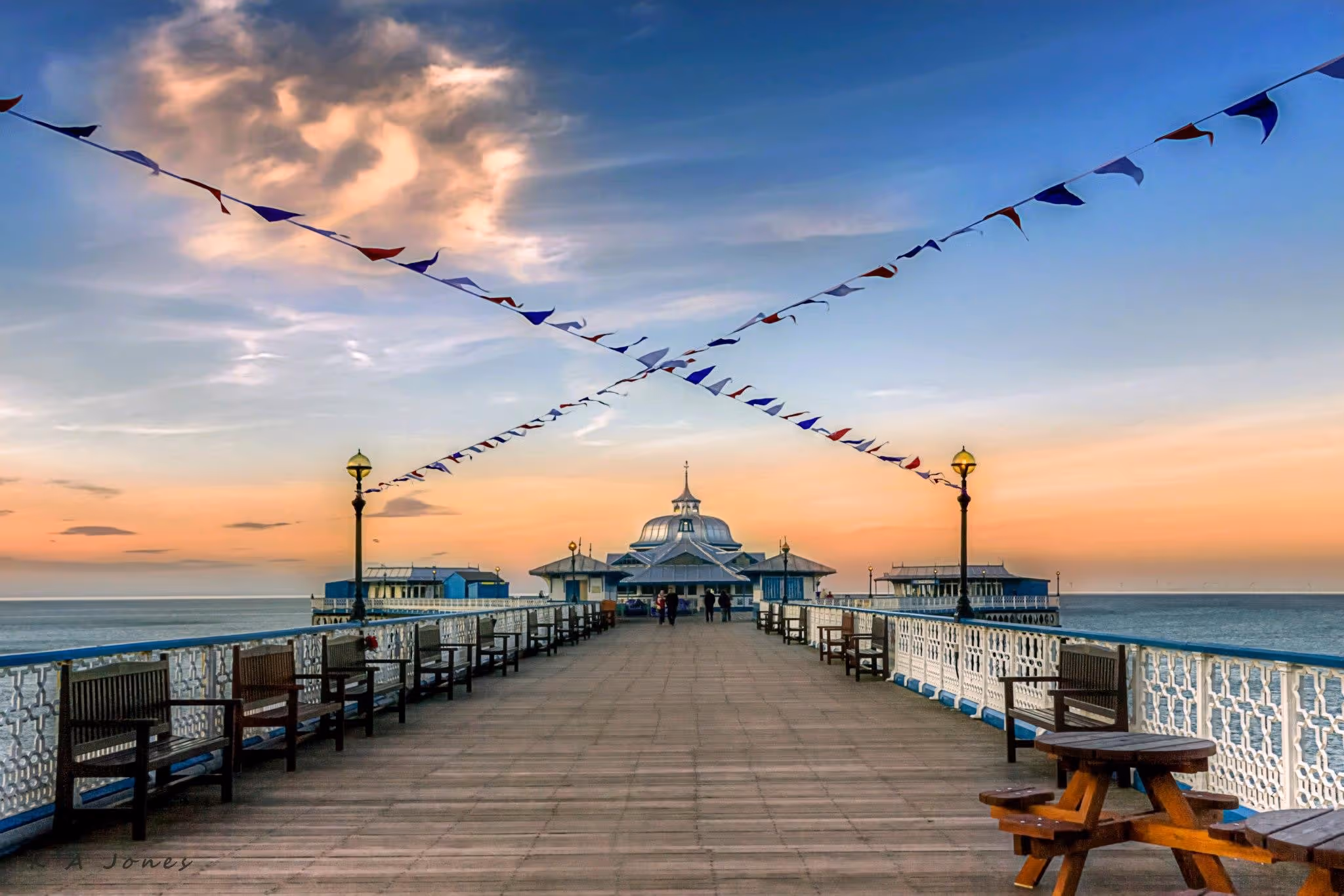 Wooden pier with benches and lamp posts, decorated with red, white, and blue bunting under a colorful sunset sky over calm sea.