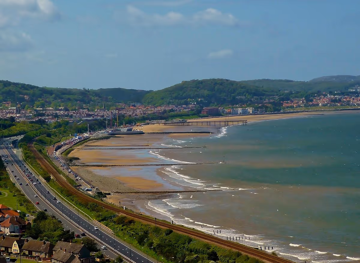 Aerial view of Colwyn Bay beach with sandy shores, green hills, coastal road, and residential area under a partly cloudy sky.