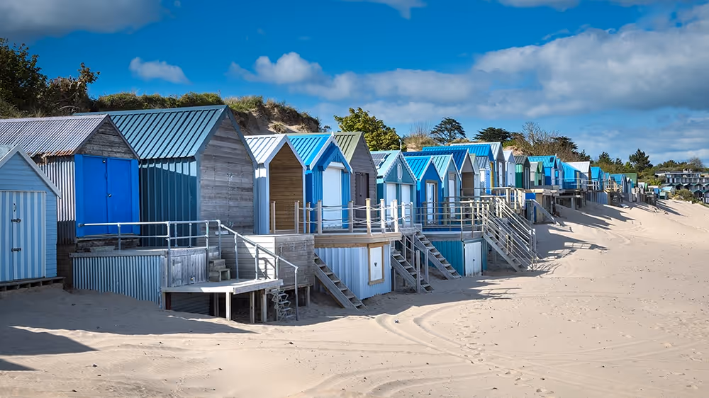 Row of colorful beach huts on a sandy shore under a blue sky with scattered clouds.