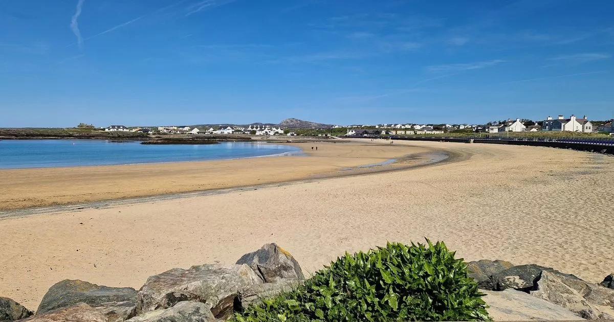 Wide sandy beach with clear blue sky, calm ocean waters, rocky foreground, and houses along the shoreline in the distance.
