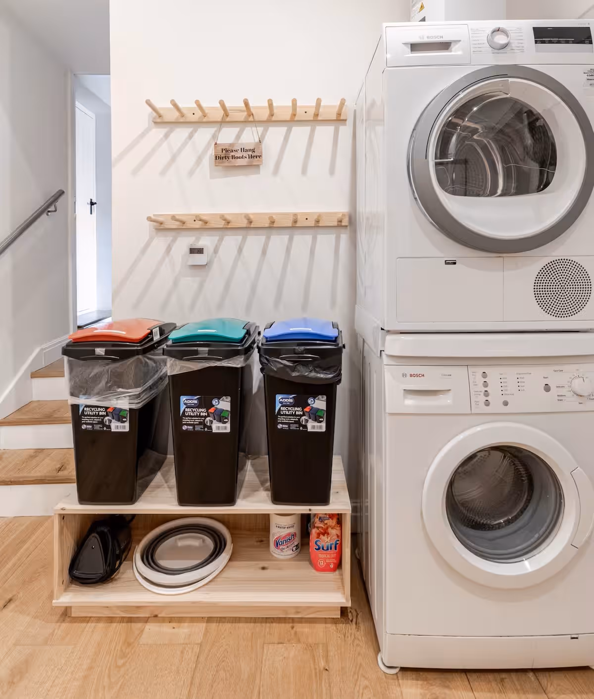 Utility Area with stacked white Bosch washer and dryer, three recycling bins with colored lids on a wooden stand, and wooden boot racks on a white wall.