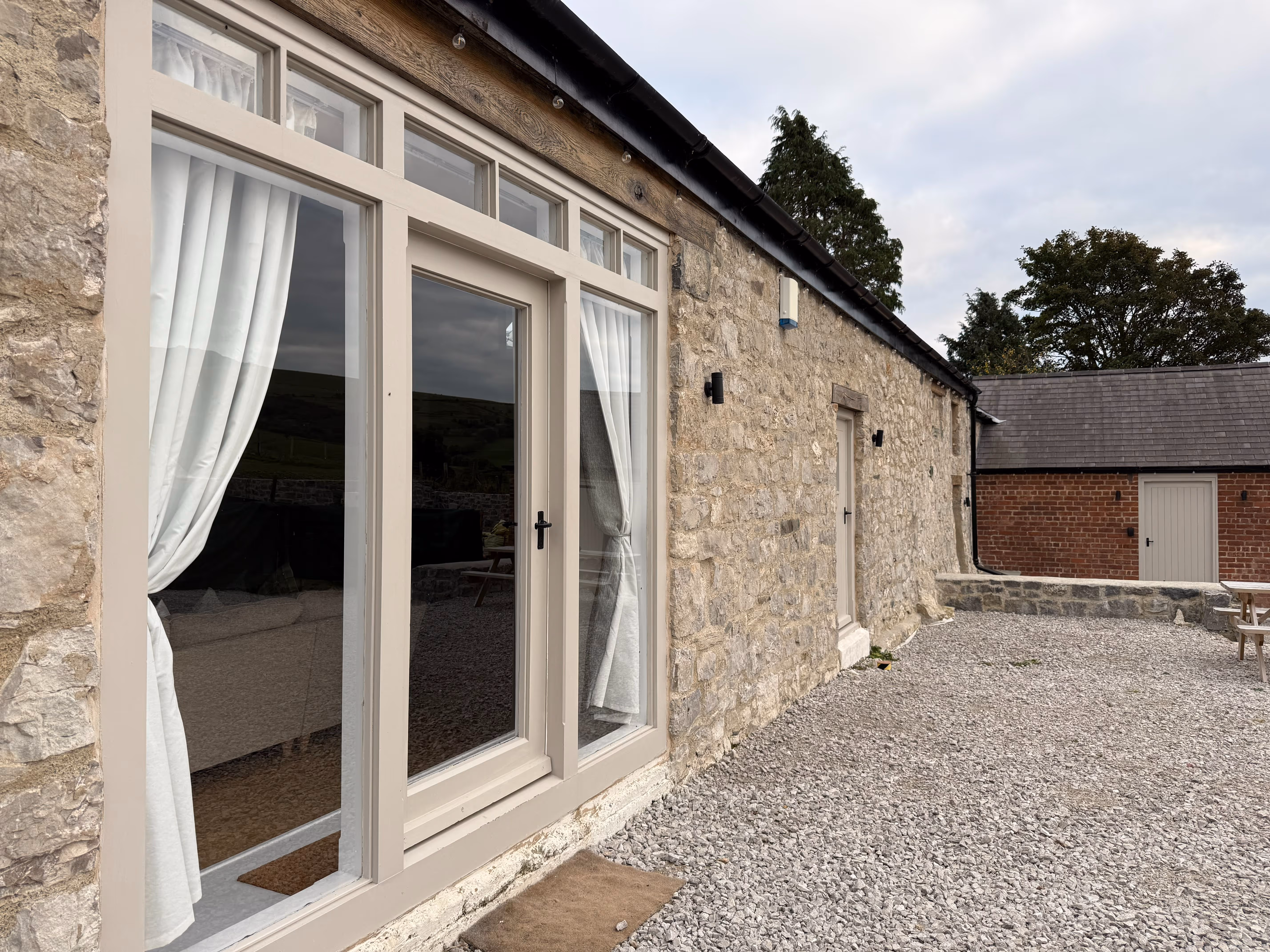 Stone cottage exterior with large glass door framed by white curtains, gravel courtyard, and secondary buildings in the background.